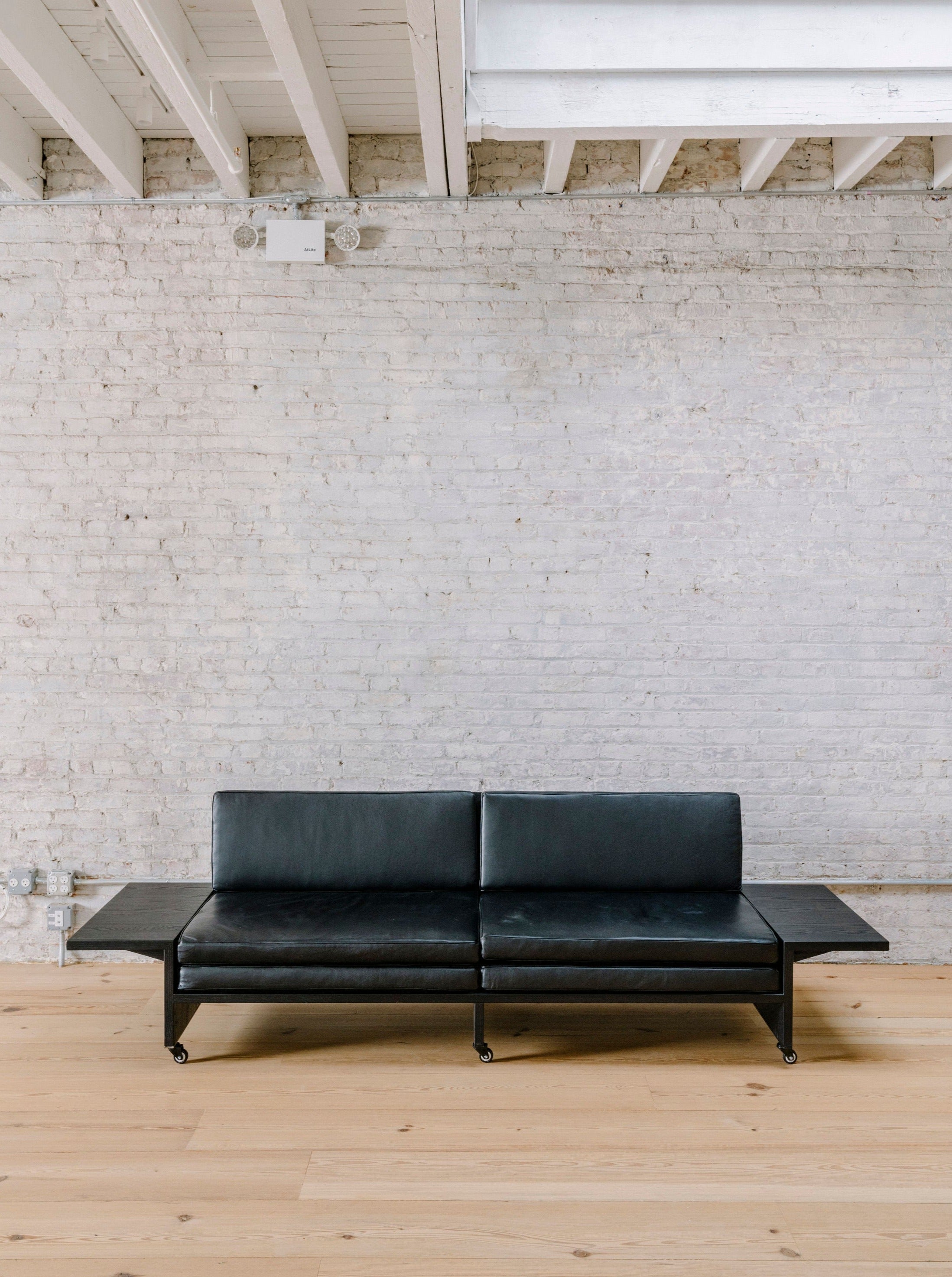A minimalist living room with a black leather modern sofa on wheels, positioned against a whitewashed brick wall. The sofa has built-in side tables, and the room features light wood flooring and exposed ceiling beams.