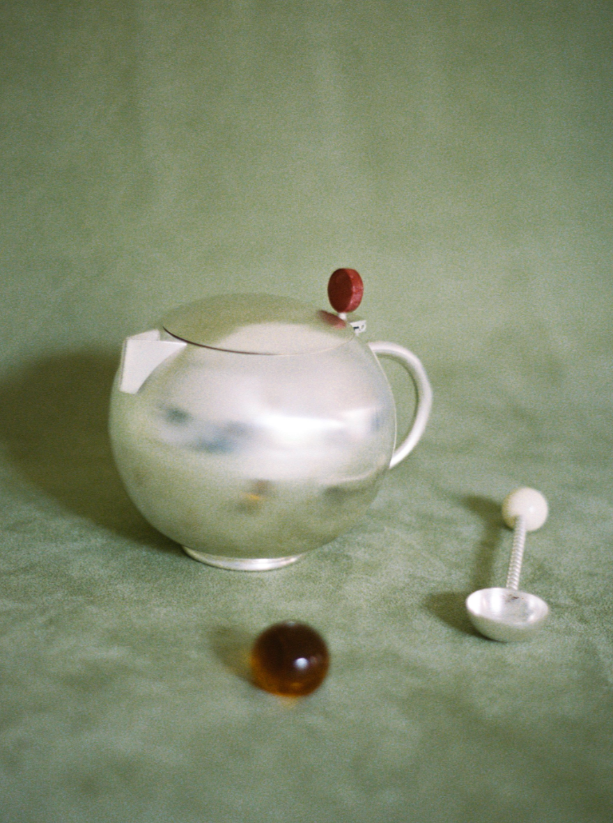 A reflective silver teapot with a red bead on the lid and a silver teaspoon, placed on a textured green surface, accompanied by a chestnut.