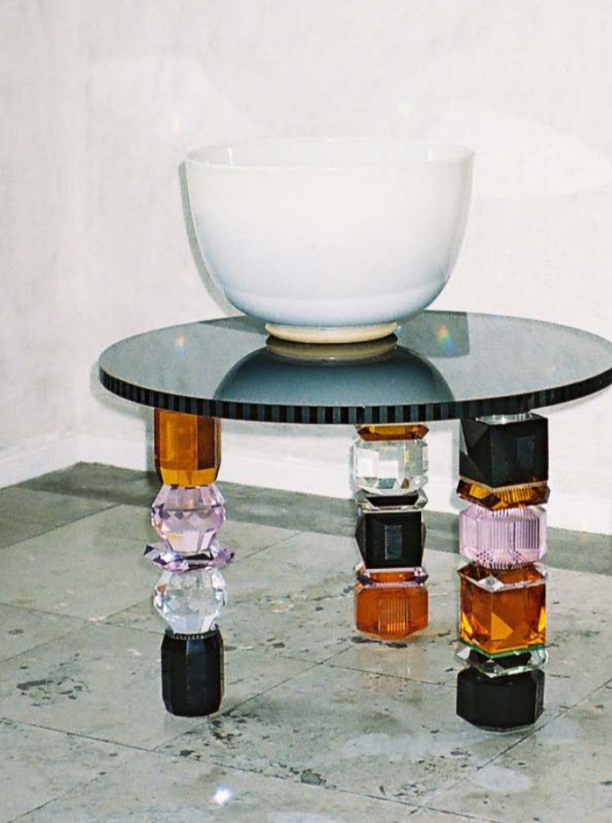 A unique side table with a glass top supported by colorful, stacked, translucent blocks of different shapes, holding a large white bowl. the setting has a tiled floor and plain white walls.