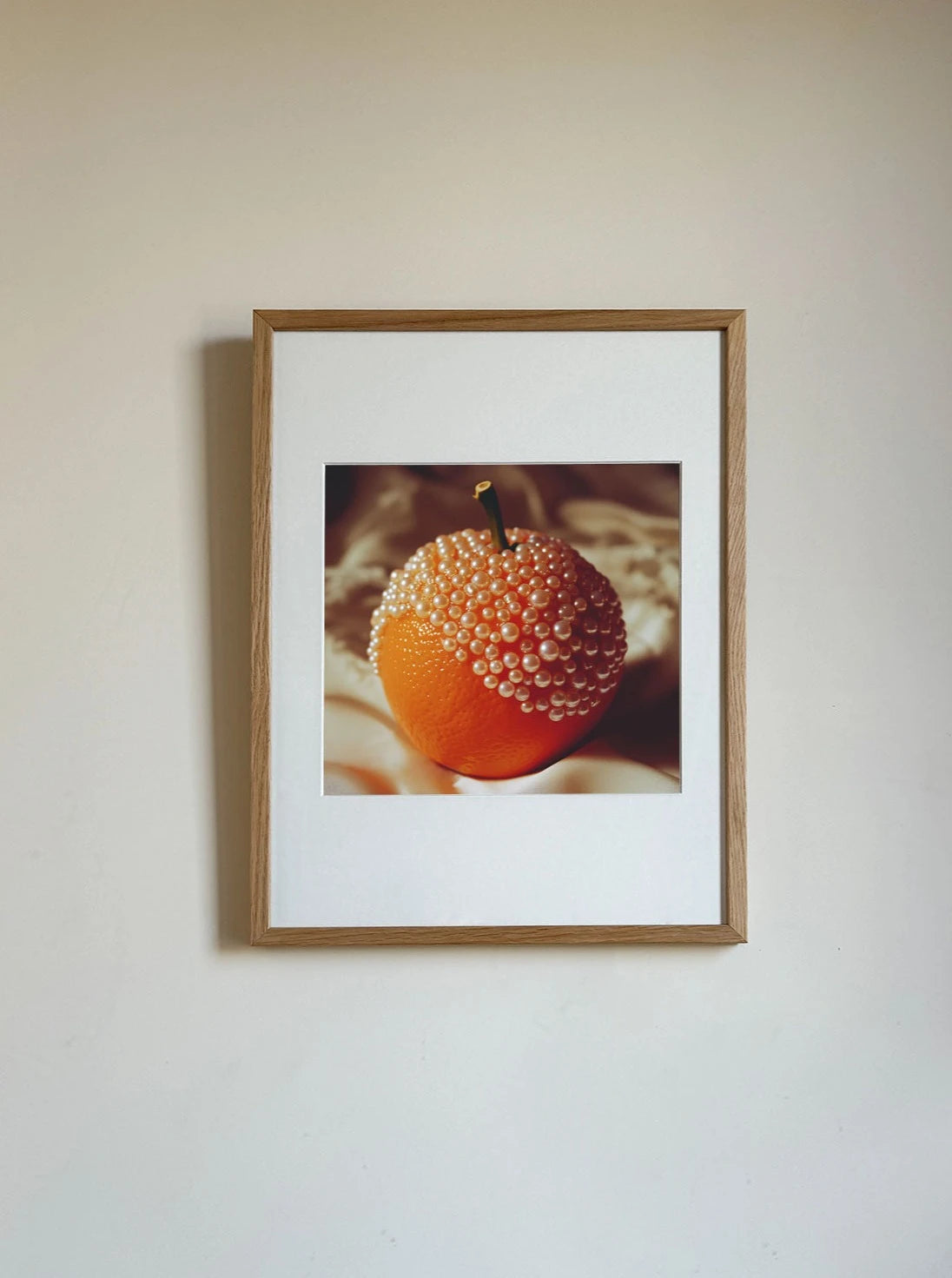 A framed photo of an orange decorated with small pearls, hanging on a beige wall. The orange has a black stem and is artistically enhanced with numerous pearls, giving it a unique and textured appearance.