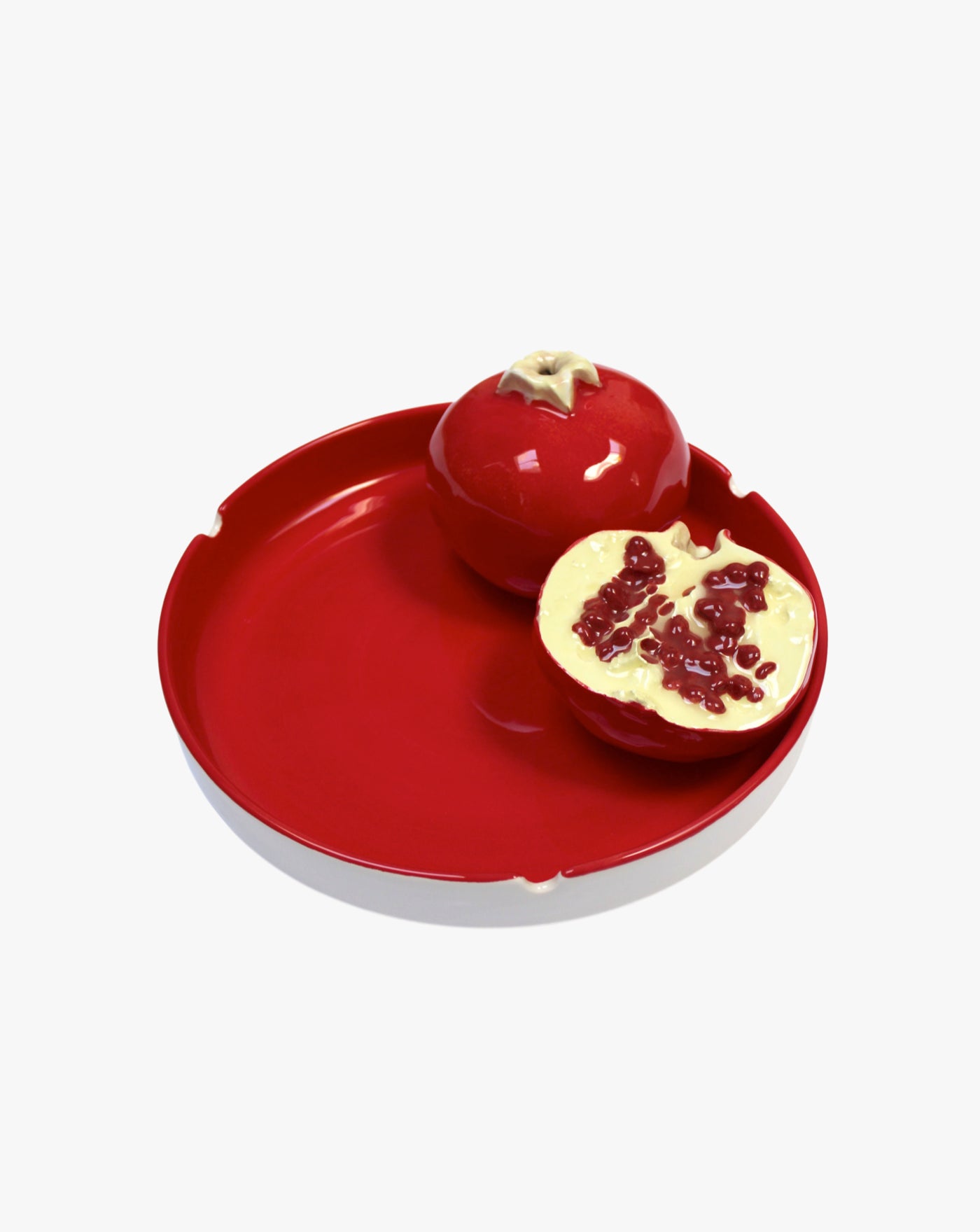 A decorative ceramic pomegranate split in half, displayed on a bright red round plate. One half is fully intact, while the other half reveals detailed seeds inside. The plate and pomegranate are placed against a white background.
