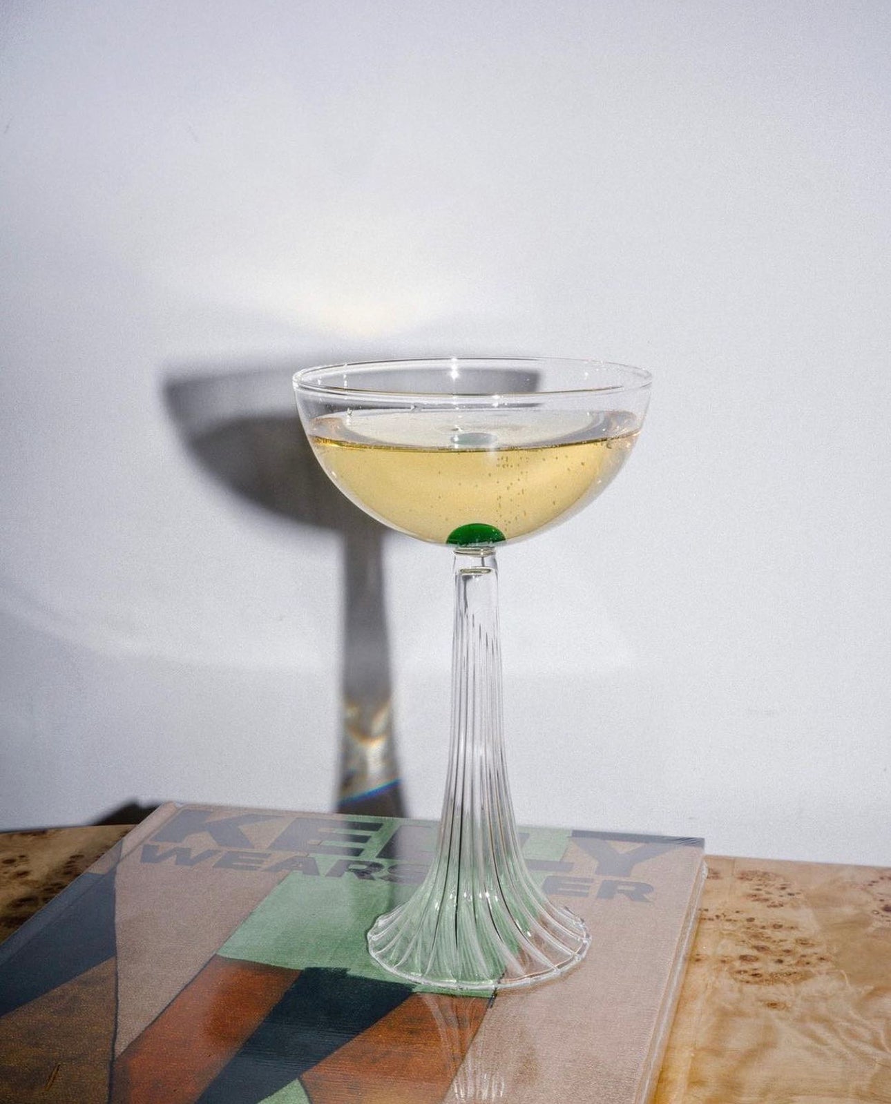A glass of white wine in an elegant, tall-stemmed glass sits on a book with abstract cover art, placed on a wooden surface. The lighting casts a soft shadow behind the glass.