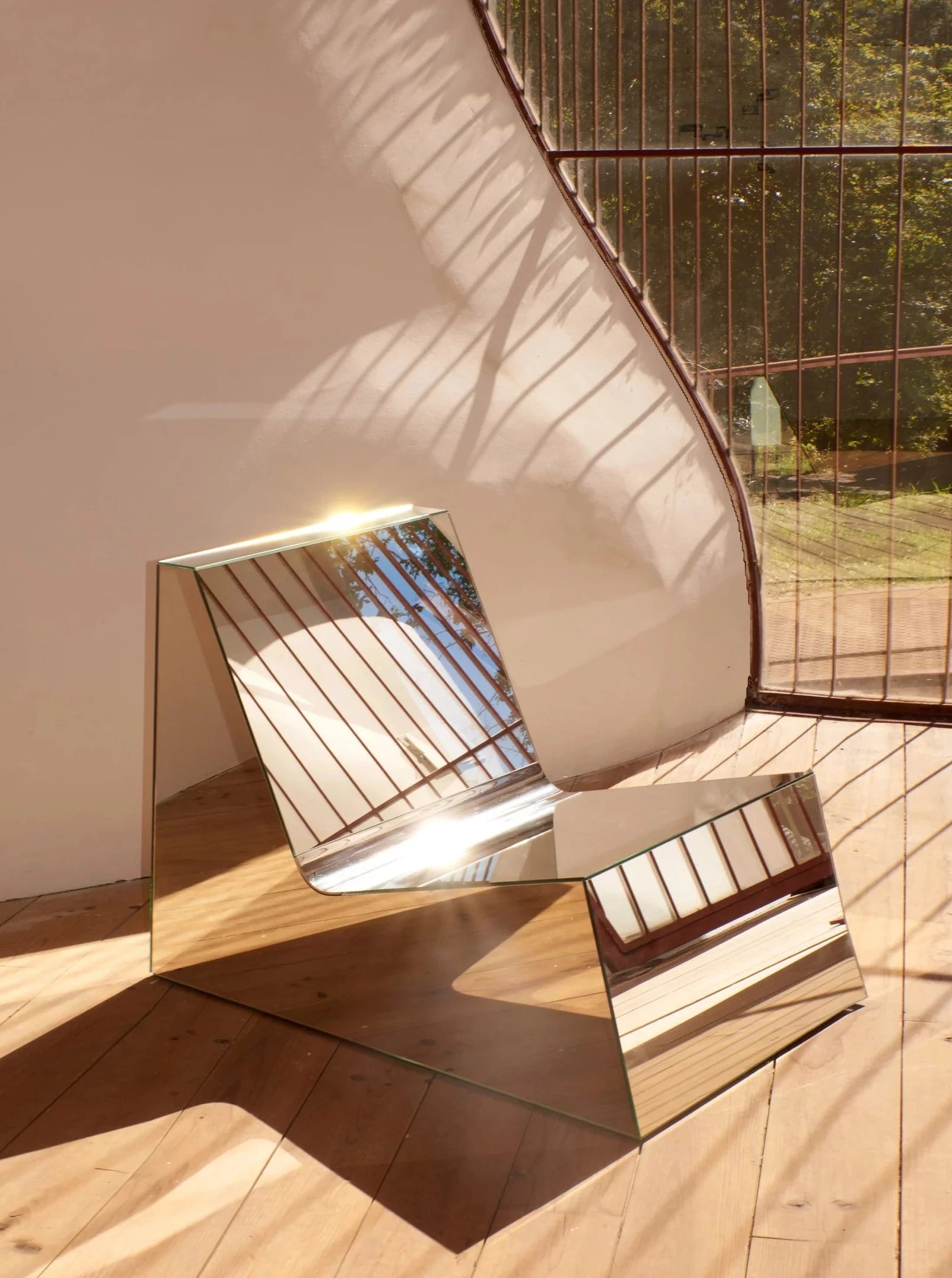 A Mirror Lounge Chair from Project 213A with a minimal shape and mirrored surface reflecting sunlight and window panes sits on a wooden floor in a sunlit room.
