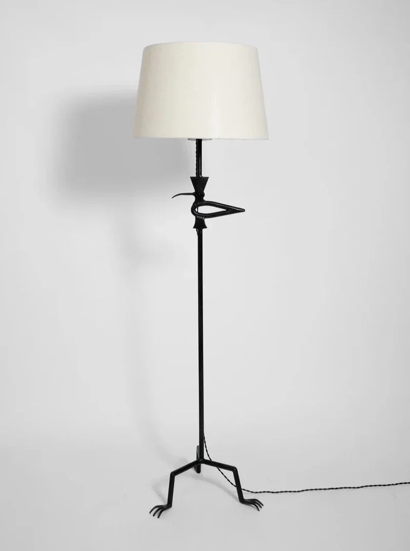 A floor lamp with a unique metal stand designed to look like bird legs, complete with feet and claws. The lamp has a white lampshade and a black base, with a power cord extending from the base.