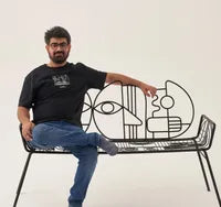 A man with a beard sits on a decorative bench with abstract line art. He's wearing a black T-shirt and blue jeans, with his legs crossed. The background is plain white.