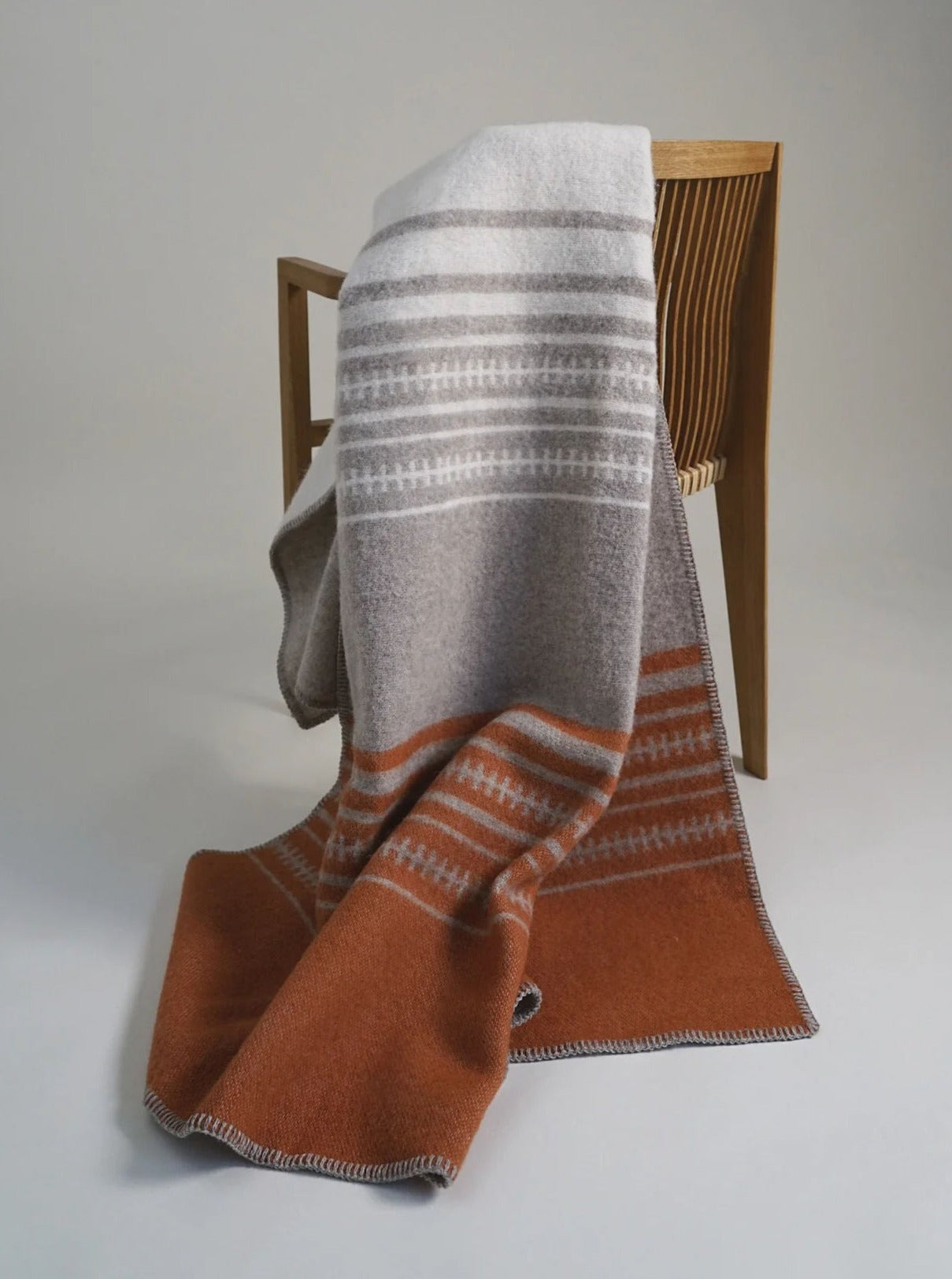 A cozy, striped Luxury Throw Blankets in shades of gray and rust hangs draped over the back of a wooden chair. The background is a plain, light-colored wall and floor.