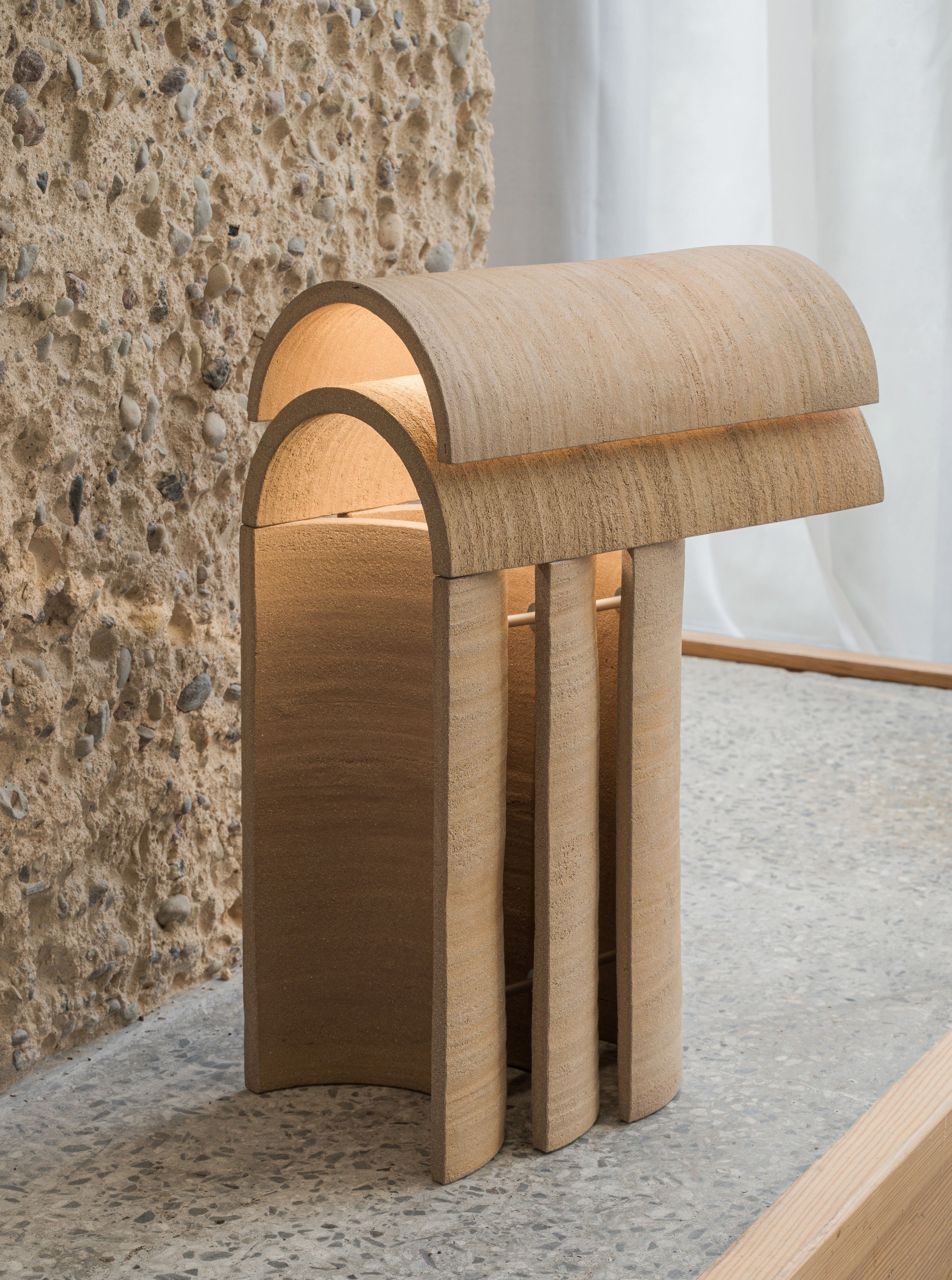 A modern wooden table lamp design with a sculptural arched design and vertical pillars, illuminated from within, placed against a stone wall and a window with a sheer curtain, Layers of Light I by Evelina Kudabaite shines beautifully.