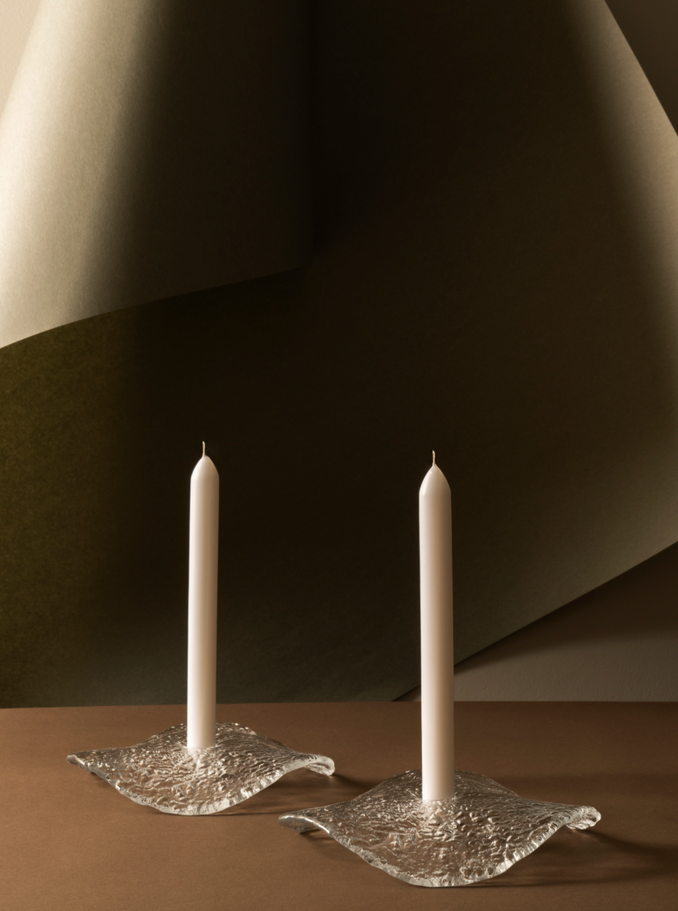 Two white taper candles standing upright in textured, clear glass holders set against a backdrop of softly draped beige and olive green fabric. The scene has a minimalist, elegant aesthetic.