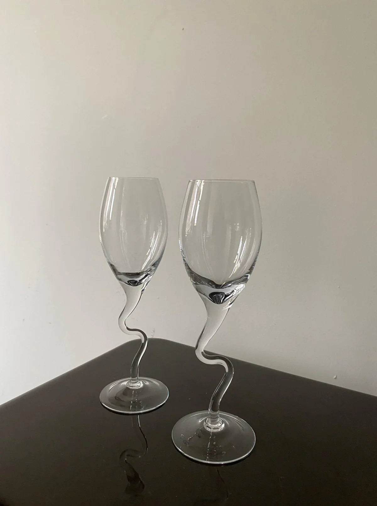 Two clear wine glasses with unique, twisted stems stand on a dark surface against a light grey wall.