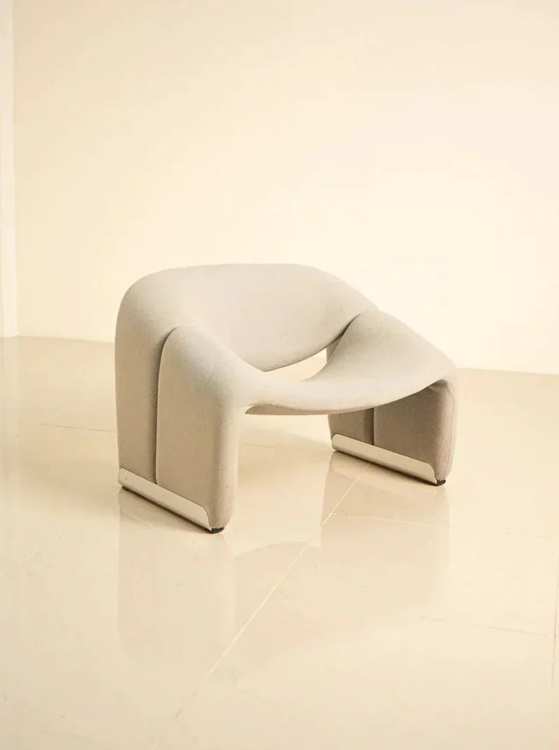 A modern, sculptural beige chair with curved lines and a minimalist design sits on a glossy cream-colored floor against a plain light wall. The chair has a unique, abstract shape with a seamless, sleek finish.