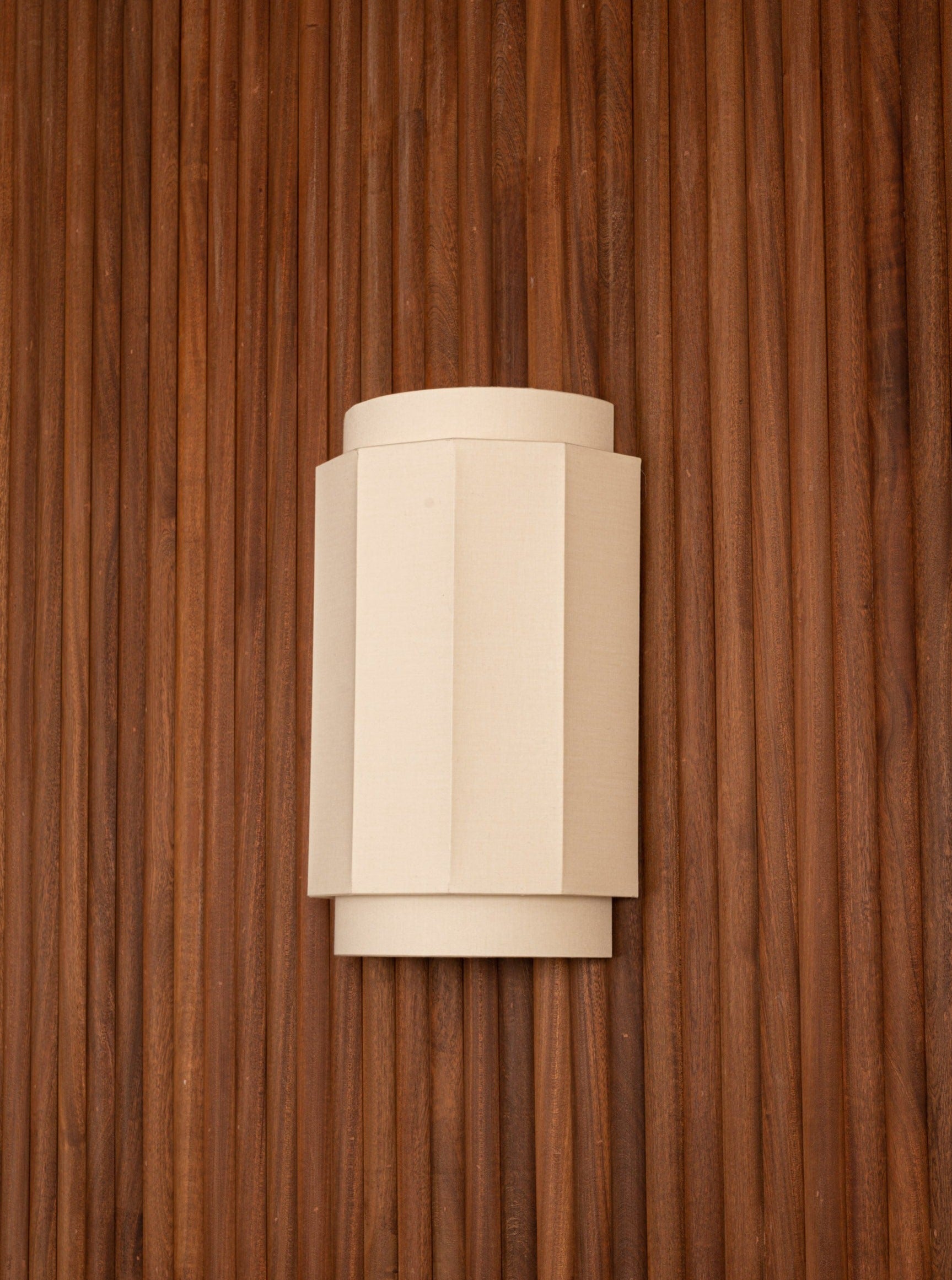 A beige wall sconce Modern Wall Lights with a geometric, faceted design is mounted on a vertical, grooved wooden panel. The wood has a rich brown color and a textured appearance, creating a striking contrast with the smooth sconce.