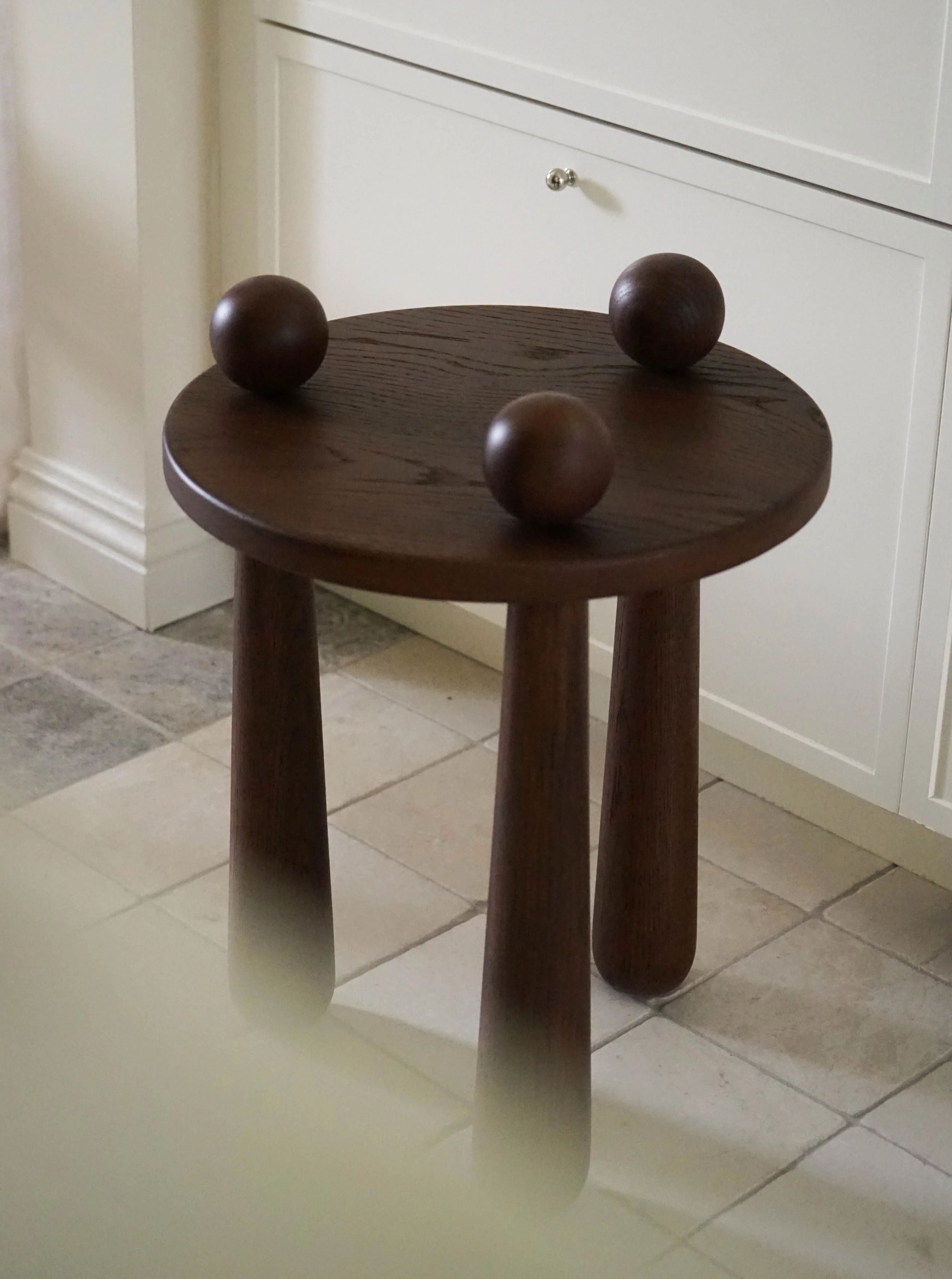 A small, round wooden interesting coffee table with three chunky legs reminiscent of a Dott Stool, Ebony stands on a tiled floor. The table has three round, dark wooden spheres placed on its surface.