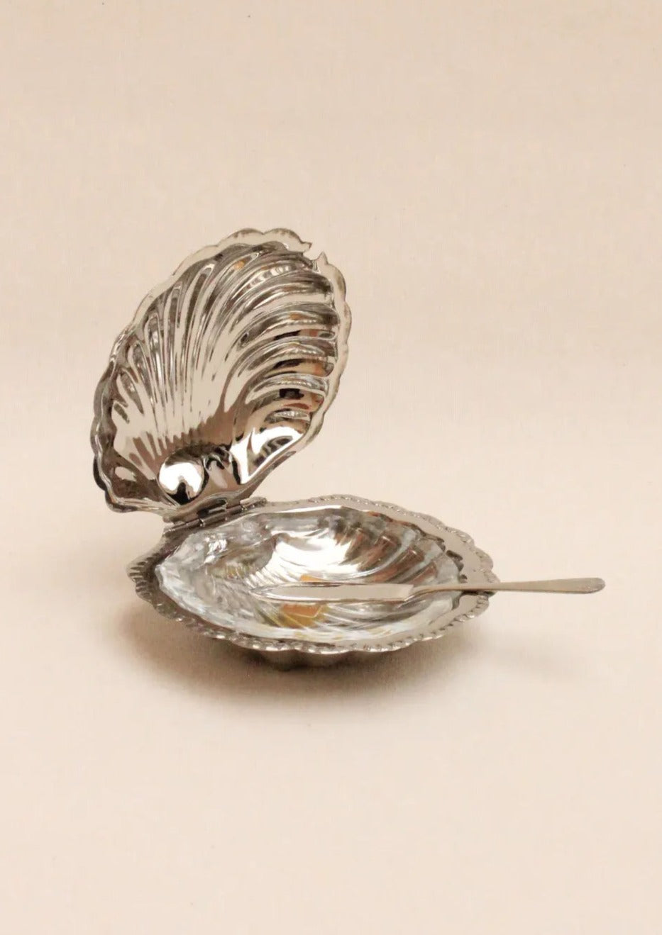 A silver clamshell-shaped dish sits open, displaying its reflective interior. A small silver spoon rests beside it, both placed on a neutral background.