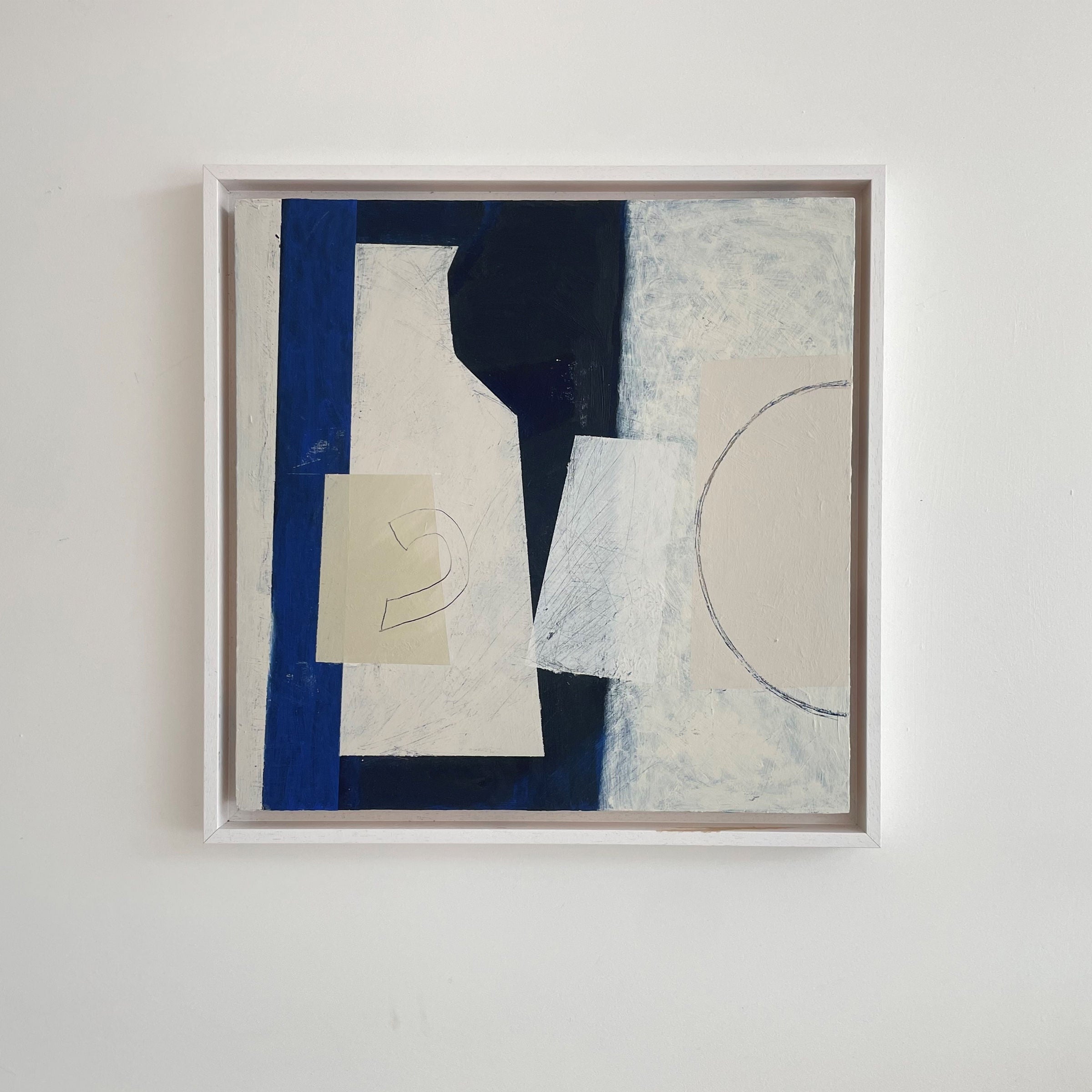 Abstract painting in a wooden frame on a white wall, featuring geometric shapes in blue, black, and white with a partial circle on the right side.