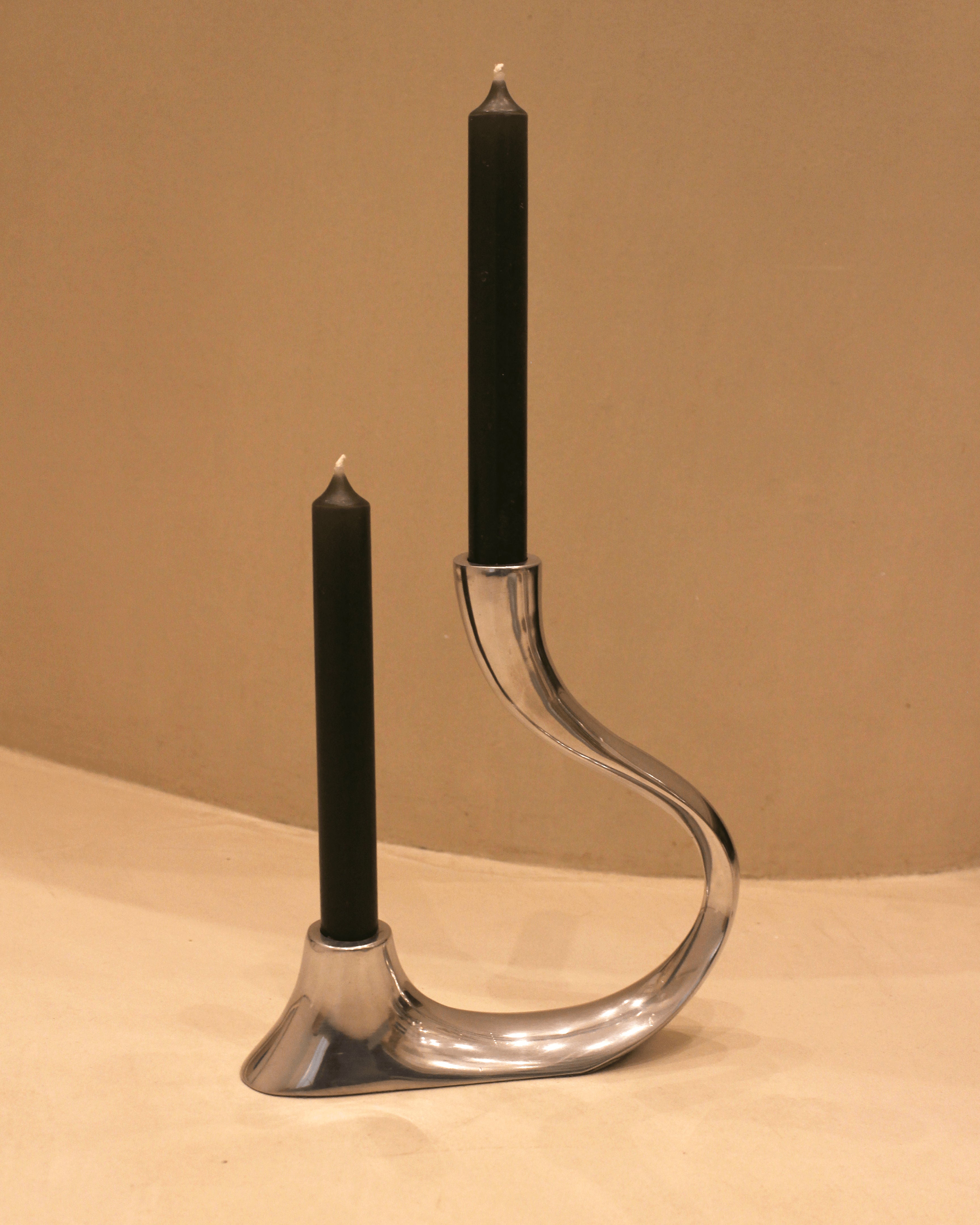 A modern, metallic, asymmetrical candle holder with two black candles. One candle is taller than the other, with the holder featuring sleek, curved lines. The background is a plain, light-colored wall.