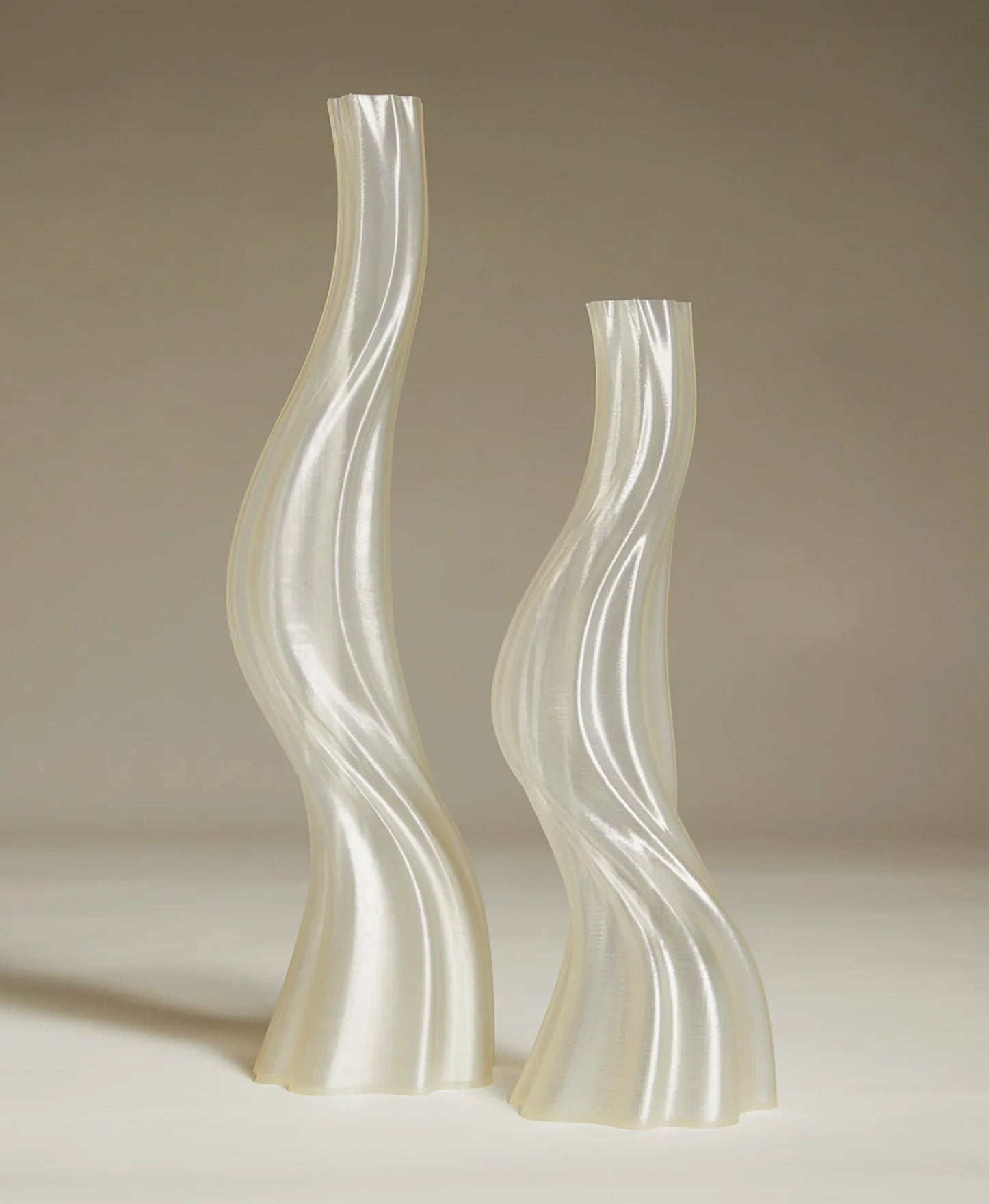 Two tall, translucent, wavy, white vases of varying heights are placed against a neutral background. The vases have an elegant, twisted design with smooth surfaces that reflect light subtly.