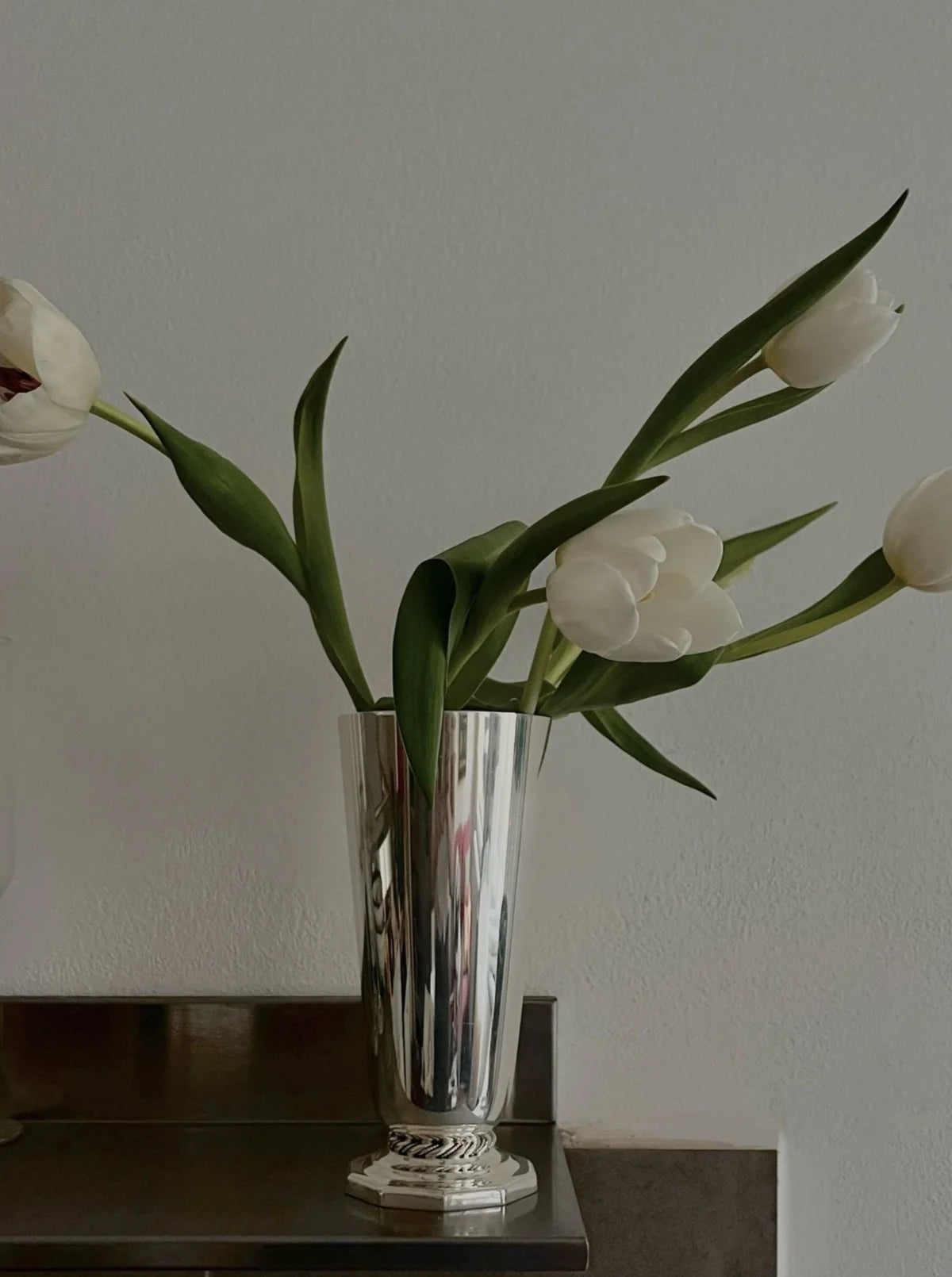 A sleek silver vase holds a minimalist arrangement of five white tulips, slightly curved, with long green stems and leaves, set against a plain white wall and dark surface.