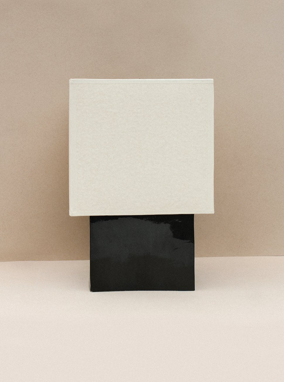 A minimalist artwork featuring two stacked geometric forms, with a textured beige square on top of a glossy black rectangle, against a neutral beige background.