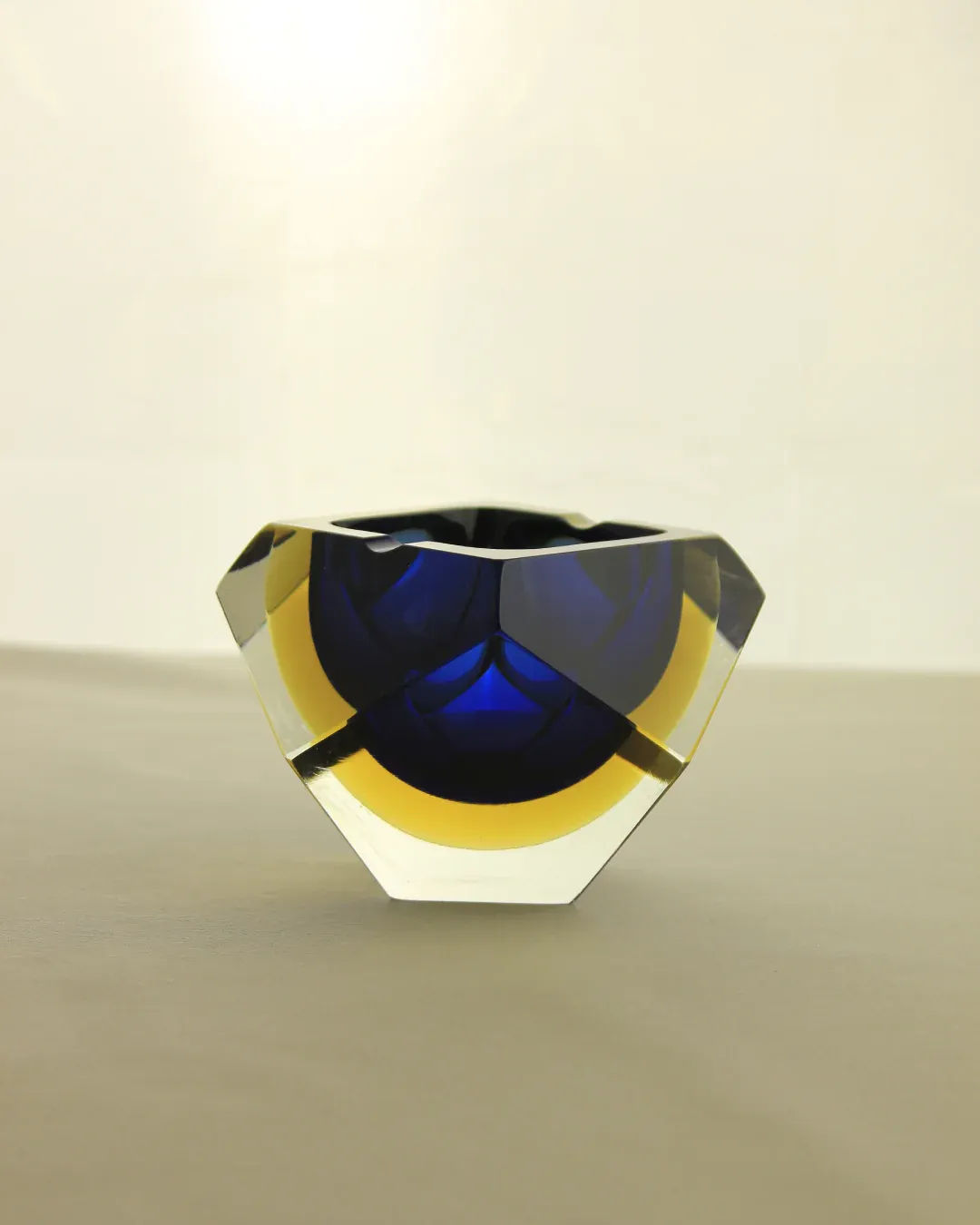 A multifaceted, transparent crystal bowl with blue and yellow accents sits on a neutral surface against a light background. The geometric design creates a modern and artistic appearance.