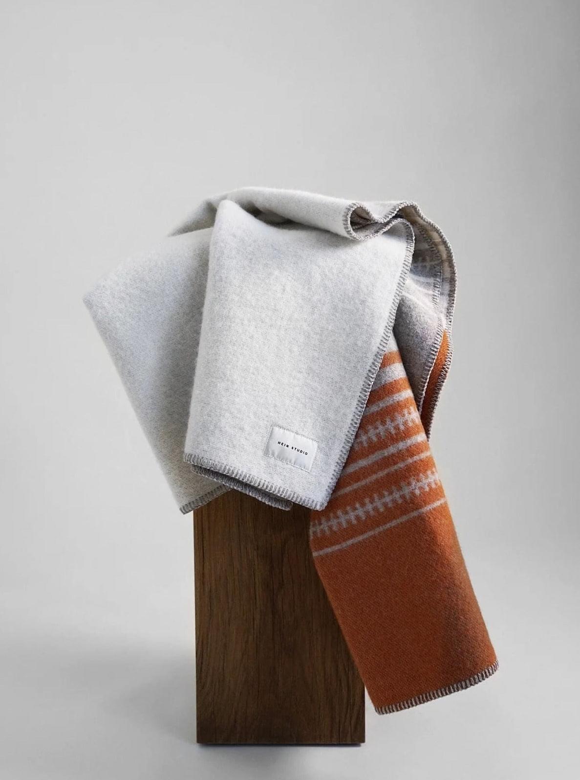 A wooden stand displaying three folded blankets; two are plain—one light gray, one darker gray—and one is patterned with Hein Studio's Aiyana Plaid - Creme/Orange.