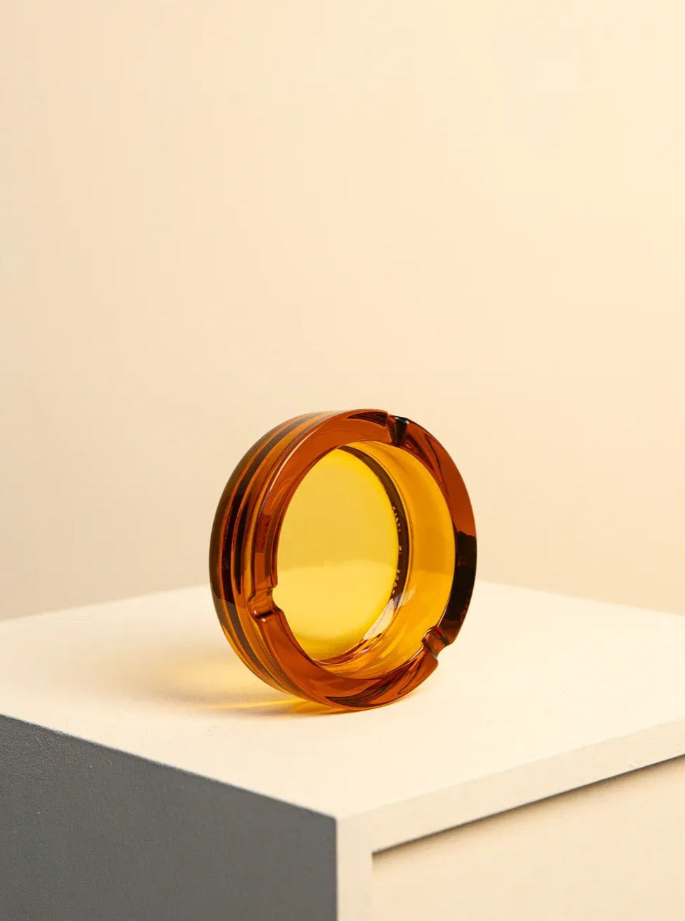 A round amber glass ashtray with sleek, modern design sits on the edge of a white and gray pedestal against a beige background.