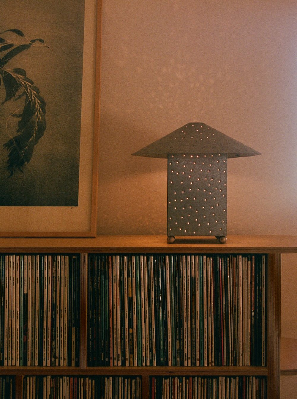 A steel, pyramid-shaped Lin Table Lamp from the MUHLY Collection with hand-punched holes casts a starry-night effect on the wall. It sits on a wooden shelf filled with numerous vinyl records. Next to the lamp is a framed picture with abstract artwork.