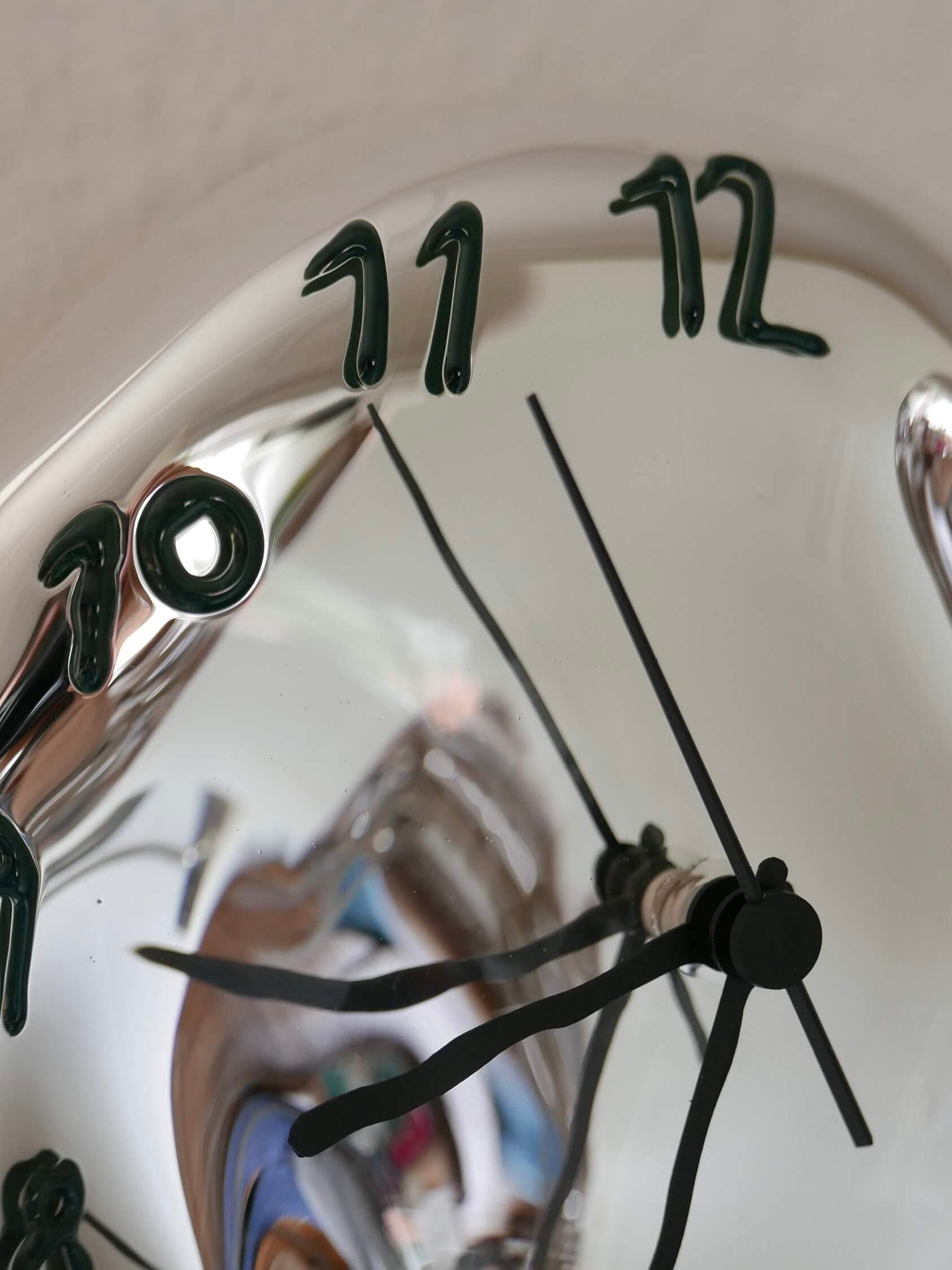 A close-up of the Silverclock - Green and Black, a handcrafted wall clock by Silje Lindrup in Denmark, features a shiny reflective surface. The numbers 10, 11, and 12 appear stretched and warped. The black clock hands are bent and twisted, adding to the melted appearance of the clock face that contrasts with its reliable timepiece origin.