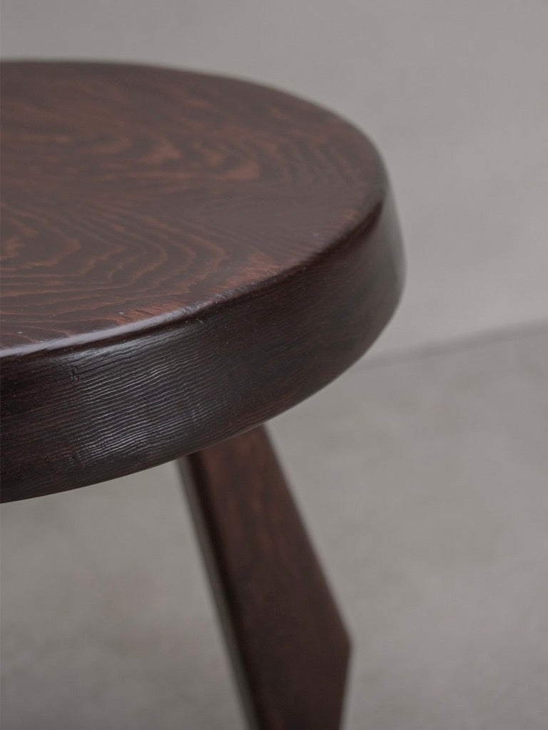 A round wooden tabletop with visible grain patterns evokes the charm of the Bicci de' Medici Charlotte Perriand - Sandoz Stool. The table's dark, polished finish contrasts with a single leg in the background on a light gray floor.