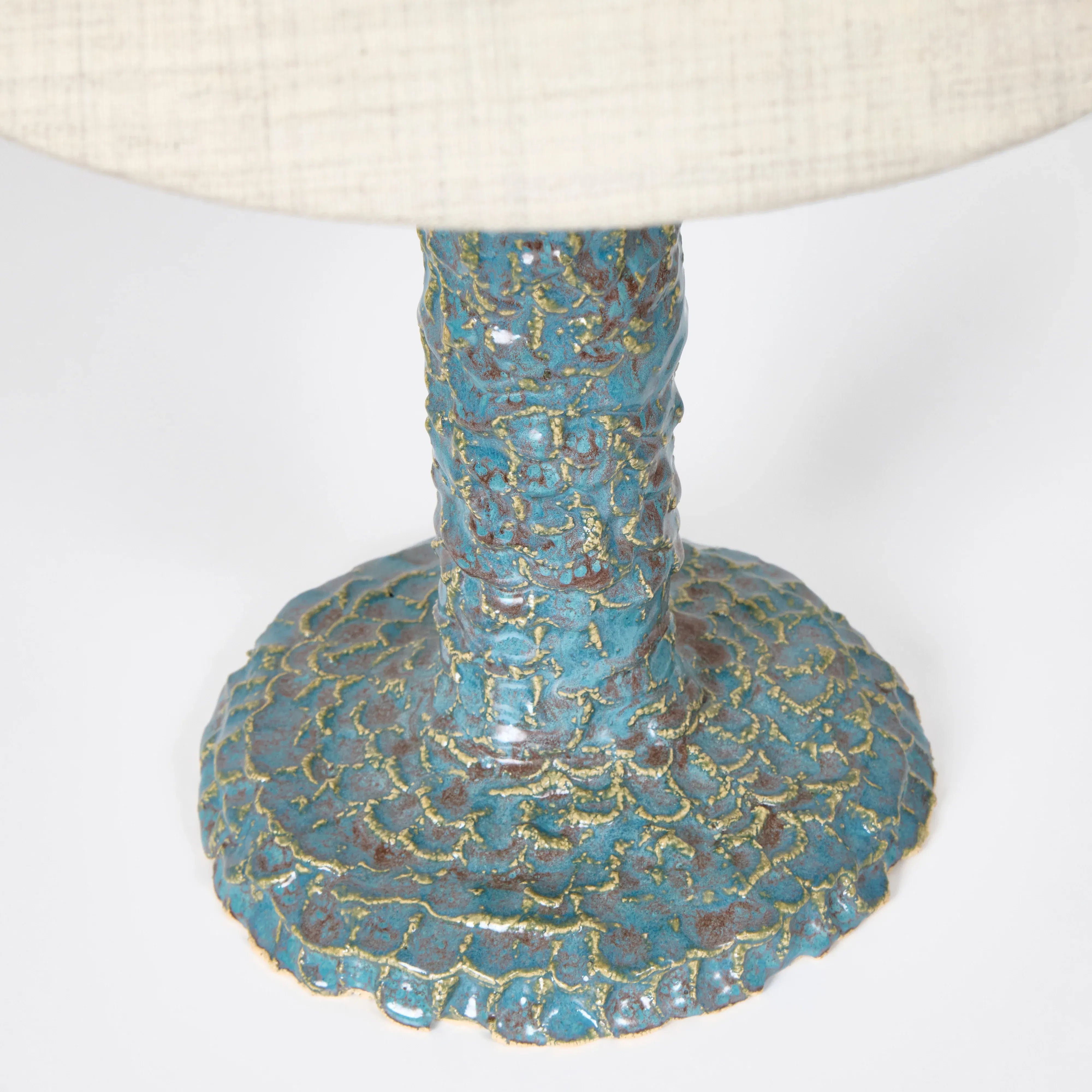 Close-up of a Project 213A Casa Bedside Light, featuring an artisanal ceramic base with a textured blue-green scaly pattern that widens at the bottom, and part of its white lampshade visible at the top.