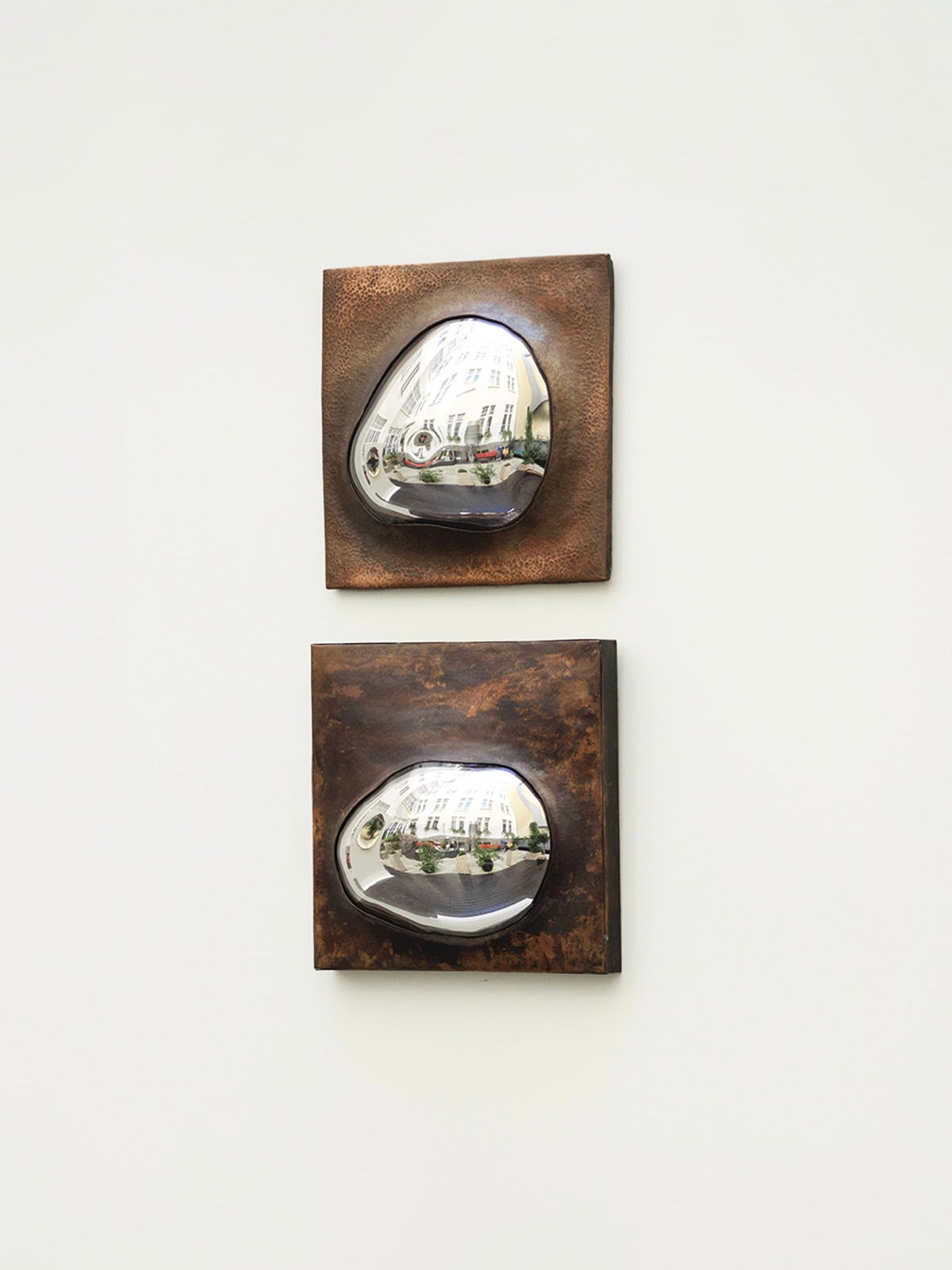 Two 'Drop Of Silver On Copper III' sculptures by HOMA, featuring reflective metallic surfaces and copper frames, are mounted on a white wall. Each sculpture is set on a square, textured brown background, reflecting a building and surrounding greenery with smooth, curved surfaces.