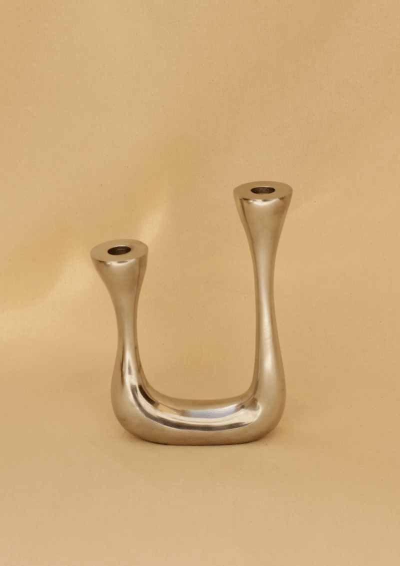 The Brutalist Candelabra by Boga Avante Shop features a modern, sculptural design with a smooth U-shape and two upward-facing slots for candles, showcased against a beige background to embrace minimalist aesthetics.