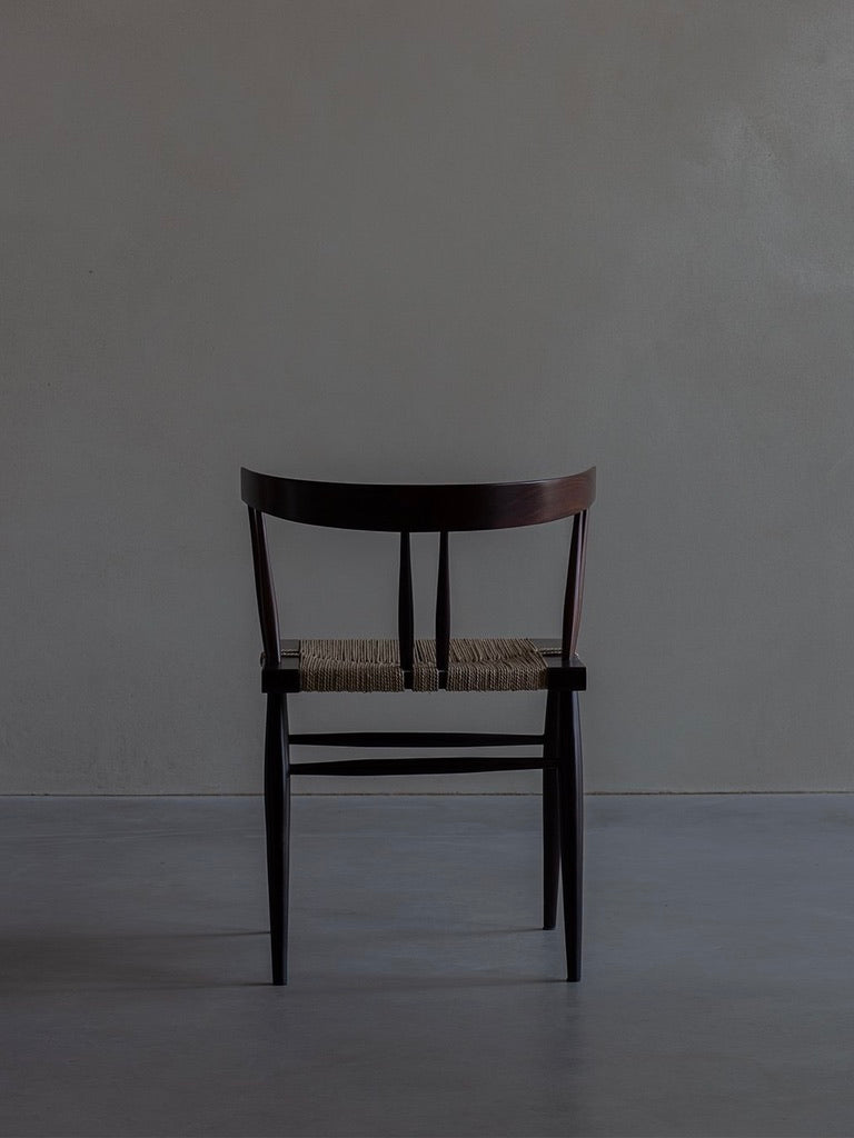 The "Manner of Nakashima - Grass Seated Chair" by Bicci de' Medici exudes mid-century allure in a minimalistic, dimly lit room. Facing a plain grey wall, its elegant dark wood and woven seat are reminiscent of George Nakashima's style on the smooth floor.