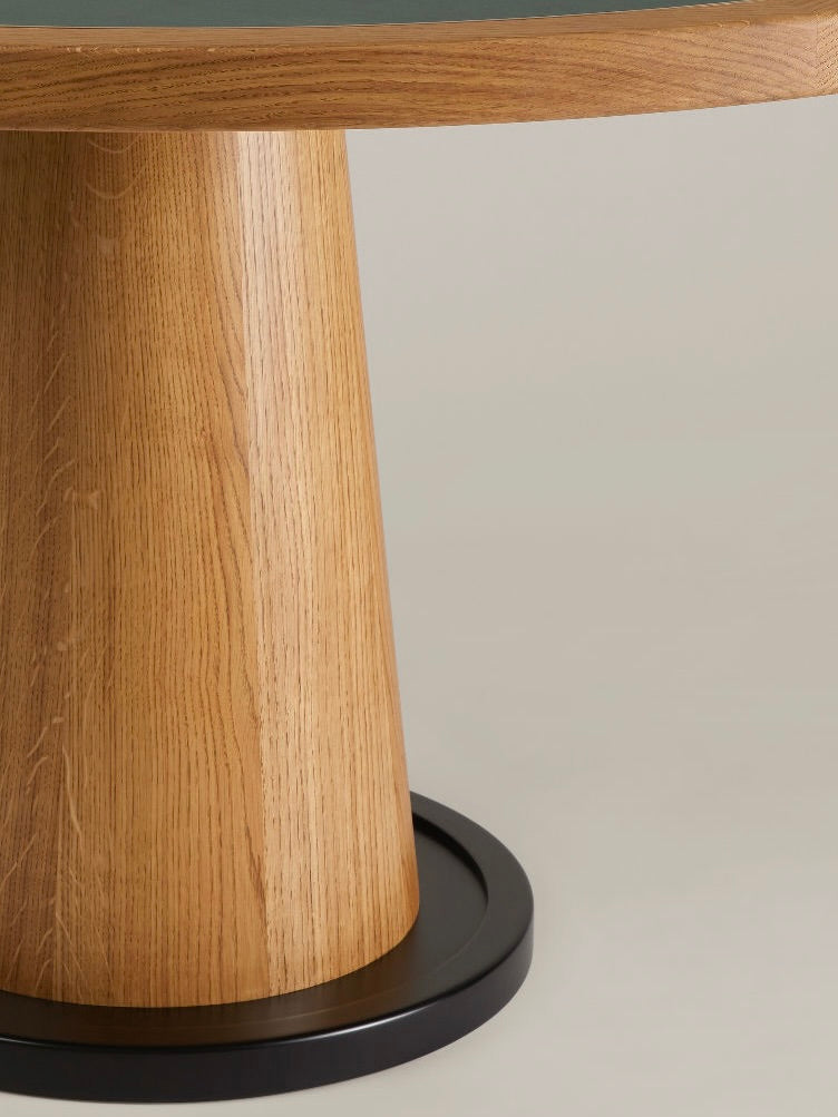 Close-up of the HAUVETTE & MADANI Jack Table, showcasing its wooden round tabletop and smooth, cylindrical base. The dark surface beautifully complements the circular black metal design of the foundation, effortlessly blending with the light-colored wood. A neutral gray background accentuates its sleek form.