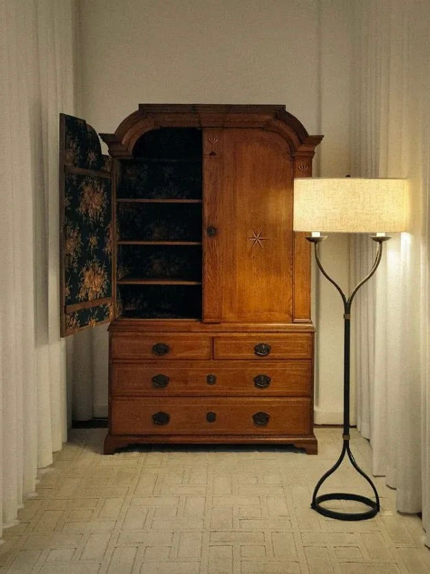 A vintage wooden wardrobe with open doors revealing shelves stands against a wall with white curtains. Next to it is the Iron Floor Lamp, 1960s by Barracuda Interiors, featuring a tall, wrought iron structure with an oval base and a beige lampshade. The scene is set on a light-colored carpeted floor.