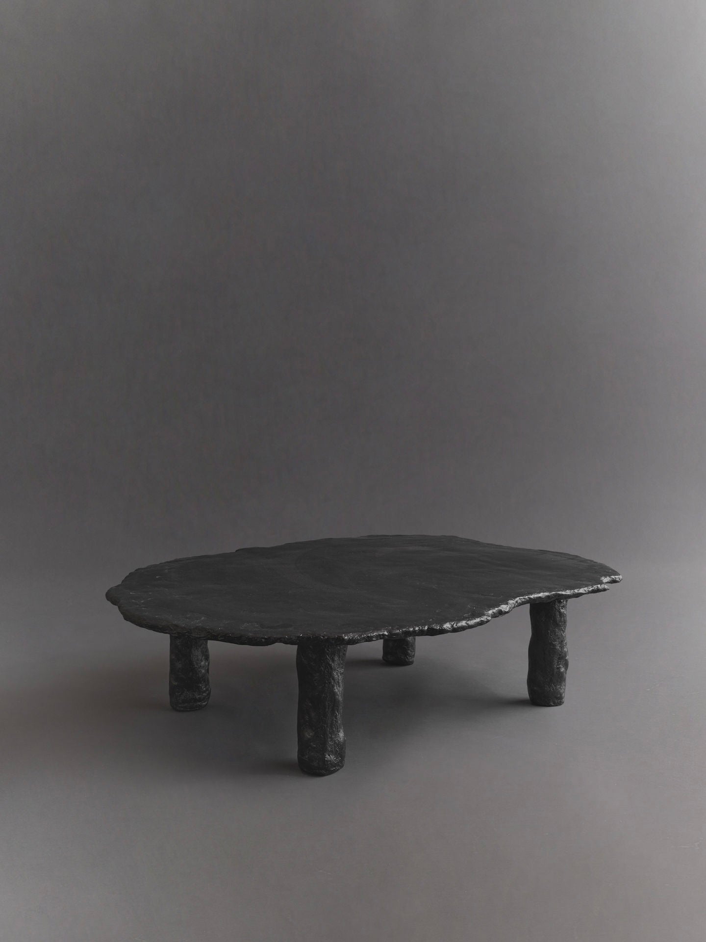 The Roca Nested Coffee Tables by Ombia Studio are handmade with a low, round design in matte black, featuring an uneven surface and organic shape. They rest on three textured cylindrical legs against a neutral gray backdrop, highlighting the tables' unique design.