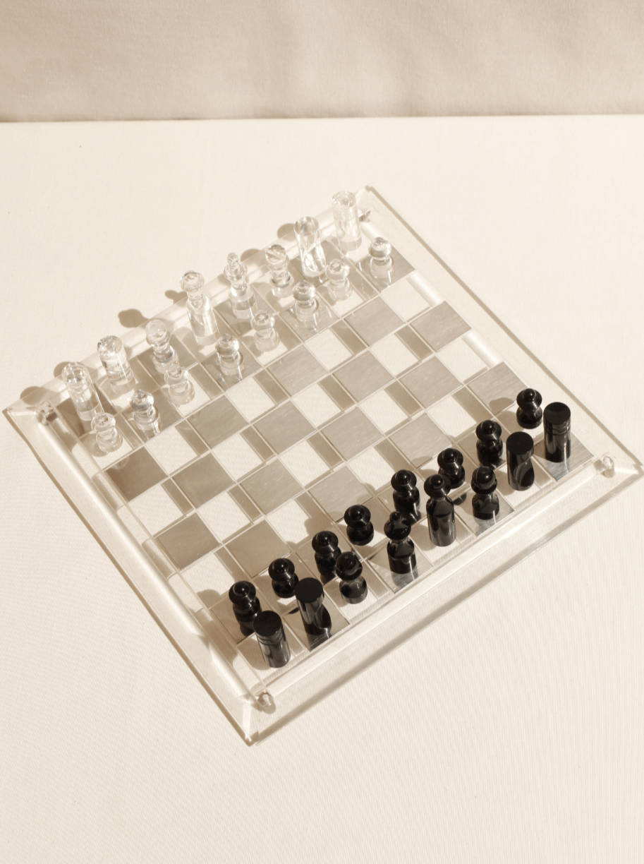 A transparent Boga Avante Shop acrylic chessboard with clear and frosted squares, featuring crystal-like clear and black chess pieces, set up and ready to play, illuminated under soft lighting.