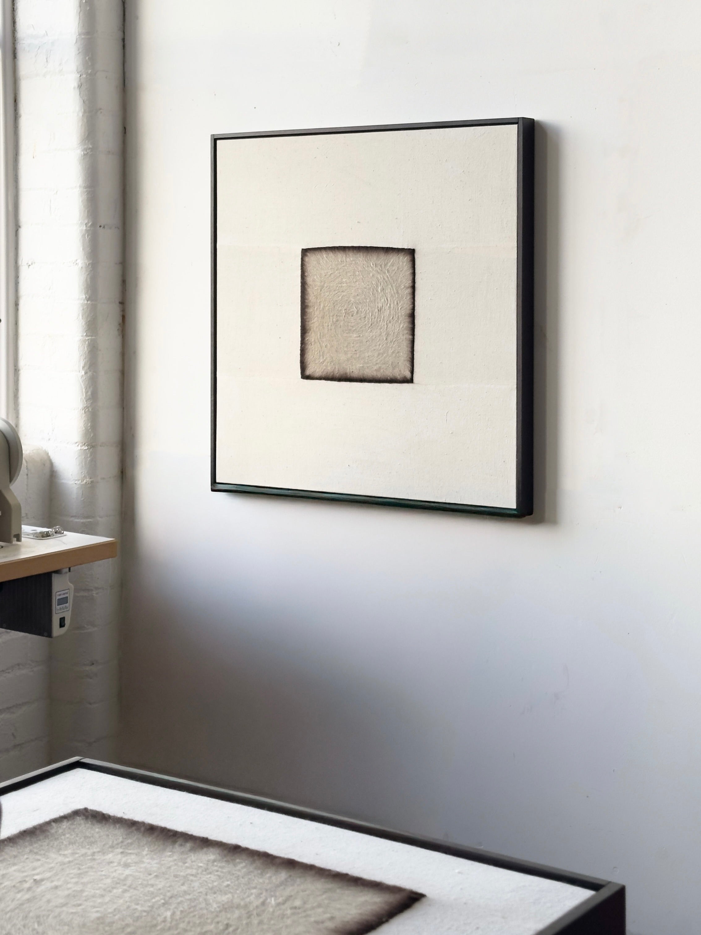 Bec Kirby's 2025 "Charred" features a centered square on a textured white background, set in a solid oak frame. It's displayed on a white wall above a worktable, with gentle light streaming in from the left window.