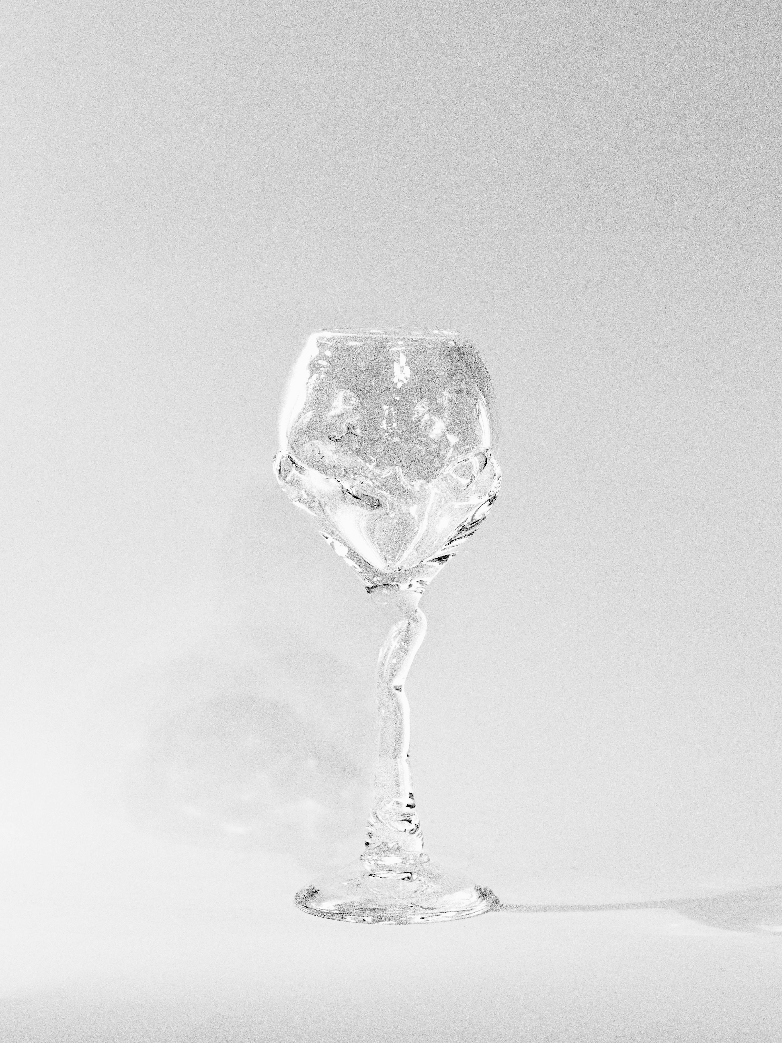 The Hand Blown Freeform Wine Glass from Szkło Studio is a clear, intricately designed piece featuring a twisted stem that stands against a plain white background. Crafted from Polish glassware, shadows cast behind it highlight its elegance and detailed features.