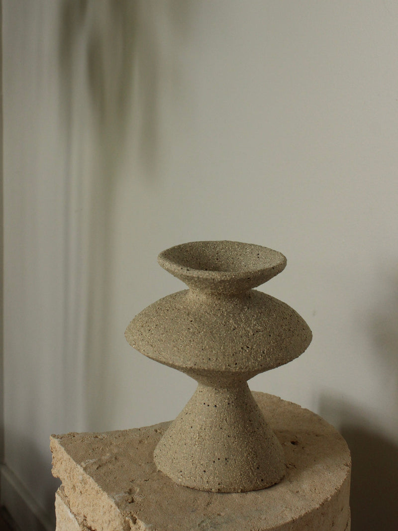 The Curve Object by Emma Lindegaard Studio, an abstract textured concrete sculpture, exudes elegance atop a stone pedestal against a neutral wall. Soft shadows enhance the minimalist composition.