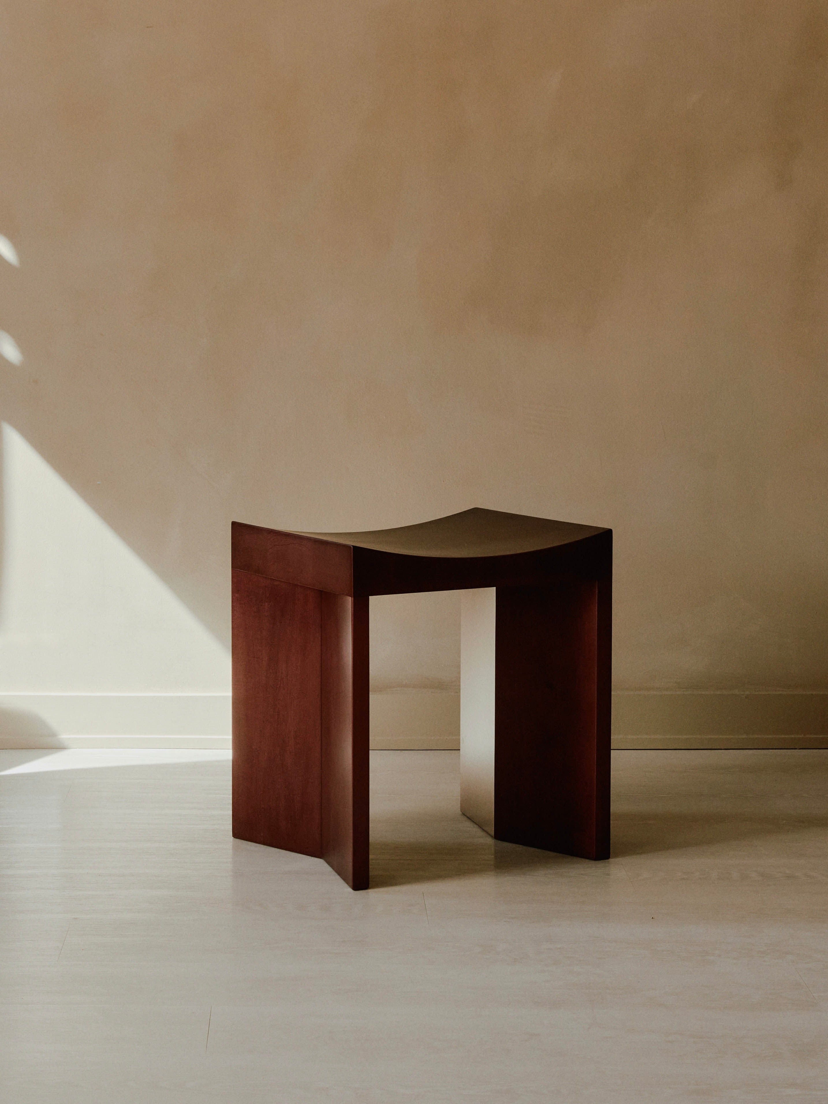 An Arc Stool by Juntos Projects, featuring a minimalist design with a curved seat, sits on a light-colored wooden floor against a beige wall. The lighting creates soft shadows, adding to the serene and simple aesthetic of the living space.