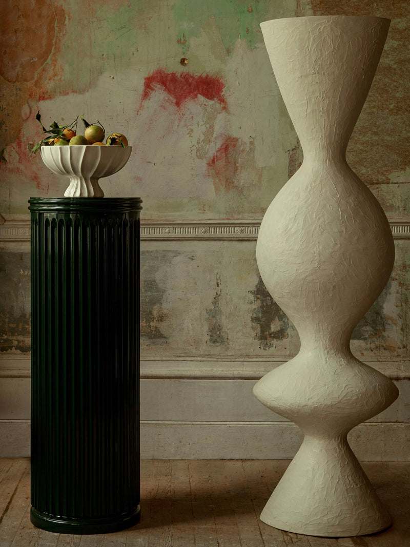 A green pedestal with a white dish, handmade in Portugal, holds fruit beside the "Venus II" Floor Lamp by Ransom & Dunn, an abstract eco-resin sculpture. The background showcases a distressed wall with faded green and red patches, contributing to a vintage ambiance.