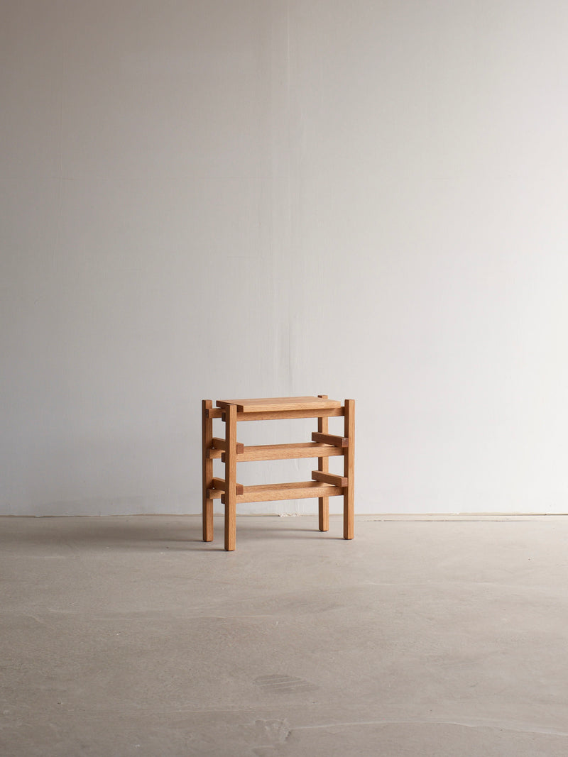 The Stool Palma by malva is a minimalist masterpiece crafted from solid oak, featuring a rectangular, three-tier design. Placed against a plain, light-colored wall on a smooth concrete floor, this stool showcases natural beauty and simple, clean lines that enhance its modern and functional aesthetic.