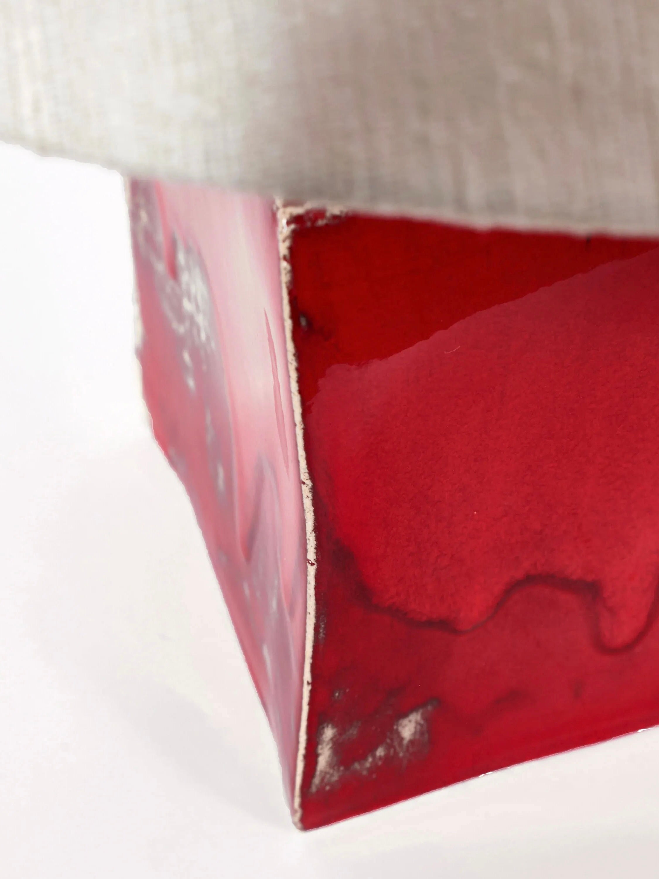 Close-up of a triangular red ceramic sculpture with uneven surfaces, drips, and rough edges reminiscent of the Arouca Table Light by Project 213A. The piece is partially covered by beige fabric at the top against a white background.