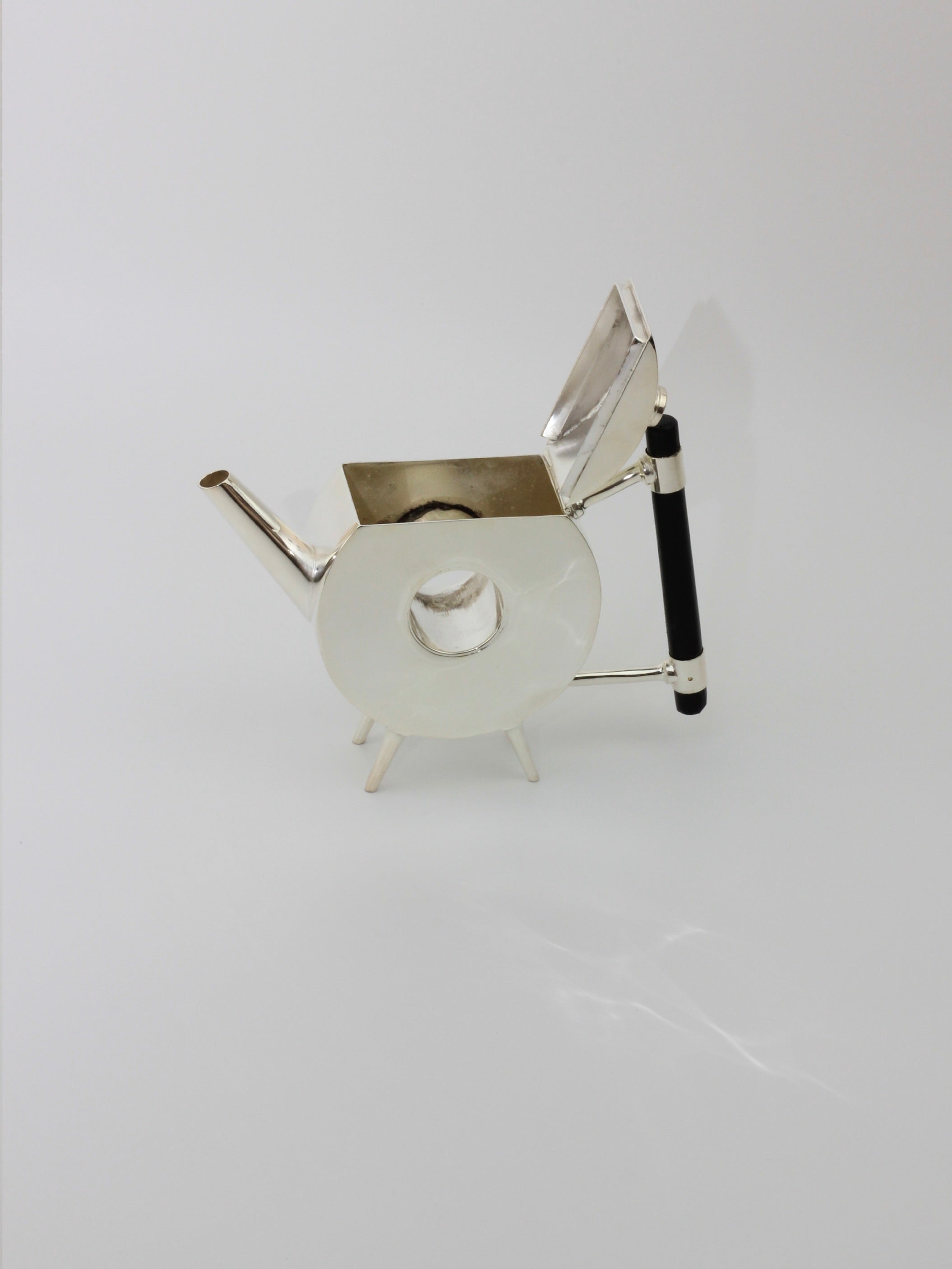 Christopher Dresser's Tea Pot from Boga Avante Shop is a modern, geometric teapot with an angular, metallic design, set against a plain white background. The silver-plated teapot features a looped circular body, a sharp-angled spout, a rectangular lid, and an ebonized wood handle. Dr. Christopher Dresser's overall design is sleek and minimalist.