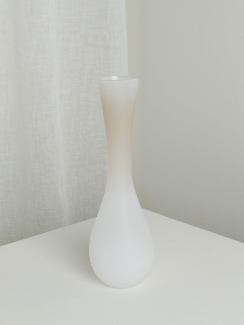 A minimalist Medium Vase in Mushroom by Verre d'Onge, featuring a frosted white finish with an elongated narrow neck and bulbous base reminiscent of its namesake, is placed on a white surface. The background showcases a light-colored fabric curtain and an off-white wall, creating a clean and serene ambiance.