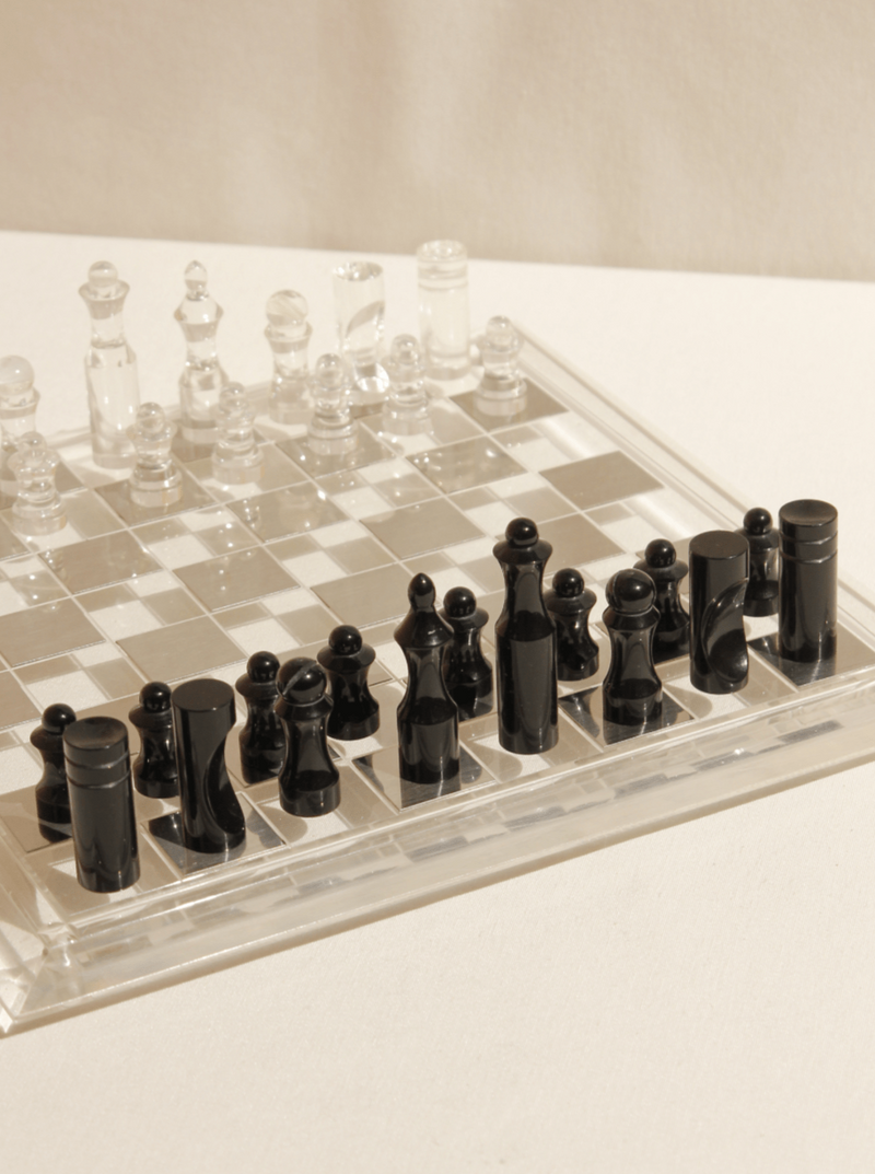 The Boga Avante Shop Acrylic Chess Board is elegantly arranged on a white surface, featuring black and clear pieces ready for play. The minimalist design enhances its elegance, casting subtle shadows.