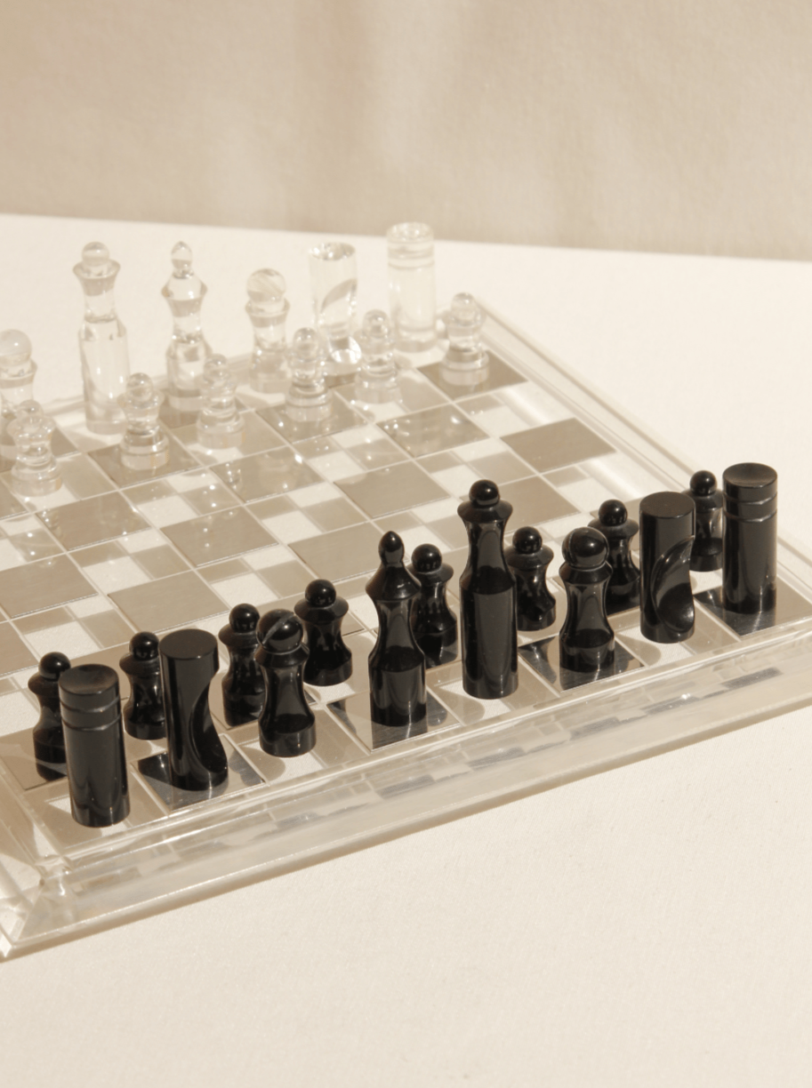 A transparent Boga Avante Shop acrylic chessboard with one set of clear and another set of black, glossy chess pieces, casting shadows on a light surface.