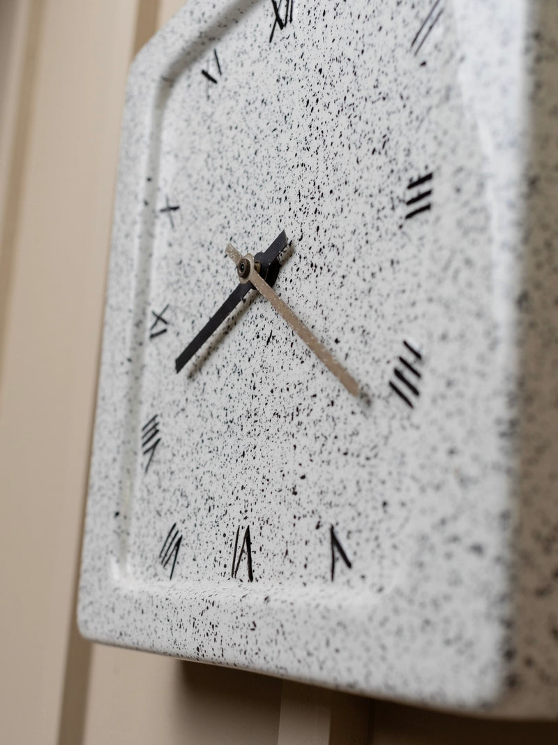 A detailed view of the Vintage Ceramic Wall Clock 1970s Italy by Bottega Jacobs showcases its speckled white face adorned with black Roman numerals. The black hour hand points to 10, while the silver minute hand rests just past 12. This delightful piece, echoing Memphis style elements, is elegantly mounted on a beige wall.
