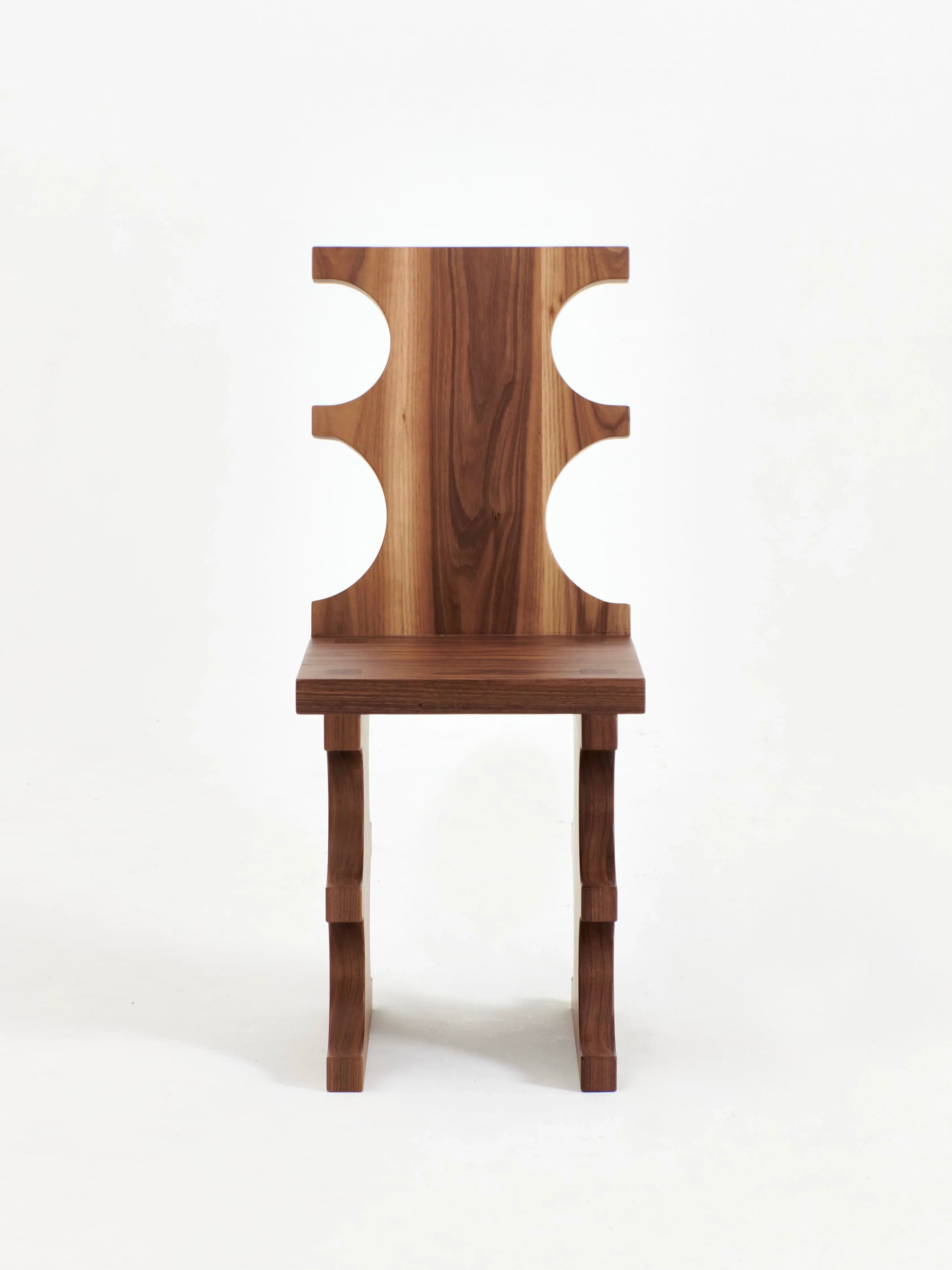 The Portugal Chair No 7 by Project 213A is seen. Crafted from solid walnut by skilled artisans, this wooden chair features a high backrest with curved cutouts on each side and a flat seat. The legs have a carved detail that complements the overall aesthetic, while the visible wood grain gives a natural look.