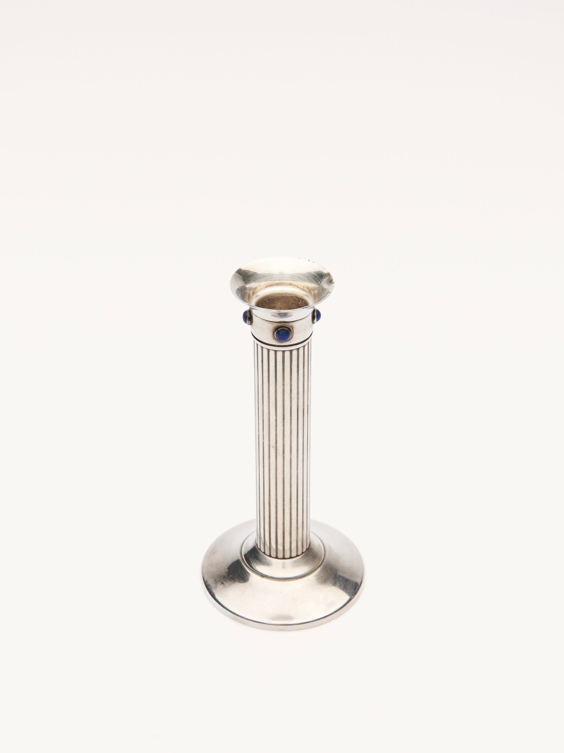 The Cartier Candle Holder by Galerie MLS boasts a sleek, modern design with a vertical ridged metallic pillar in silver-plated metal. It features a round base, a candle slot at the top, and is adorned with a small lapis lazuli stone detail against a plain white background.